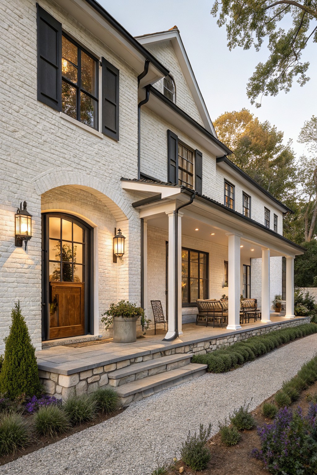 White painted brick house exterior with black shutters and windows, arched wooden front door flanked by lanterns, covered porch with columns, stone steps, and gravel path with low plantings.