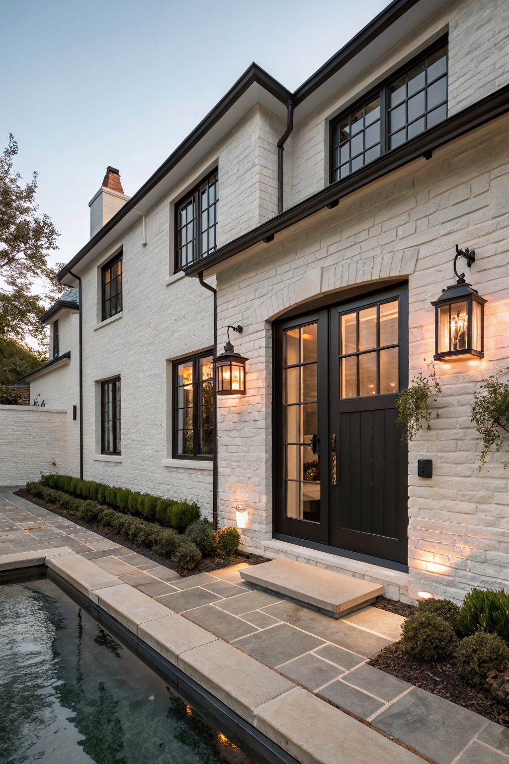 Side view of a two-story white painted brick house exterior featuring black-framed windows and double doors with an arched top, flanked by black lanterns, a stone path leading to an in-ground pool, and low boxwood hedges.