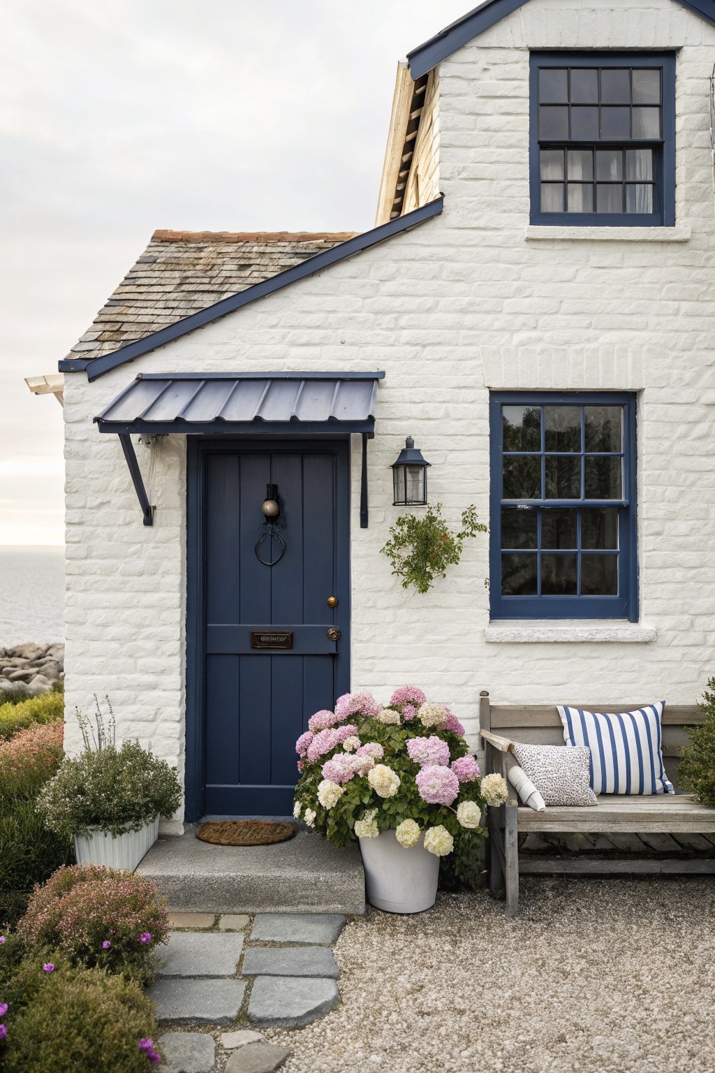 Small white painted brick house exterior featuring a navy blue front door with knocker, blue window frames, wall lanterns, potted hydrangeas and plants, wooden bench with cushions, stone steps, and gravel path near the ocean.