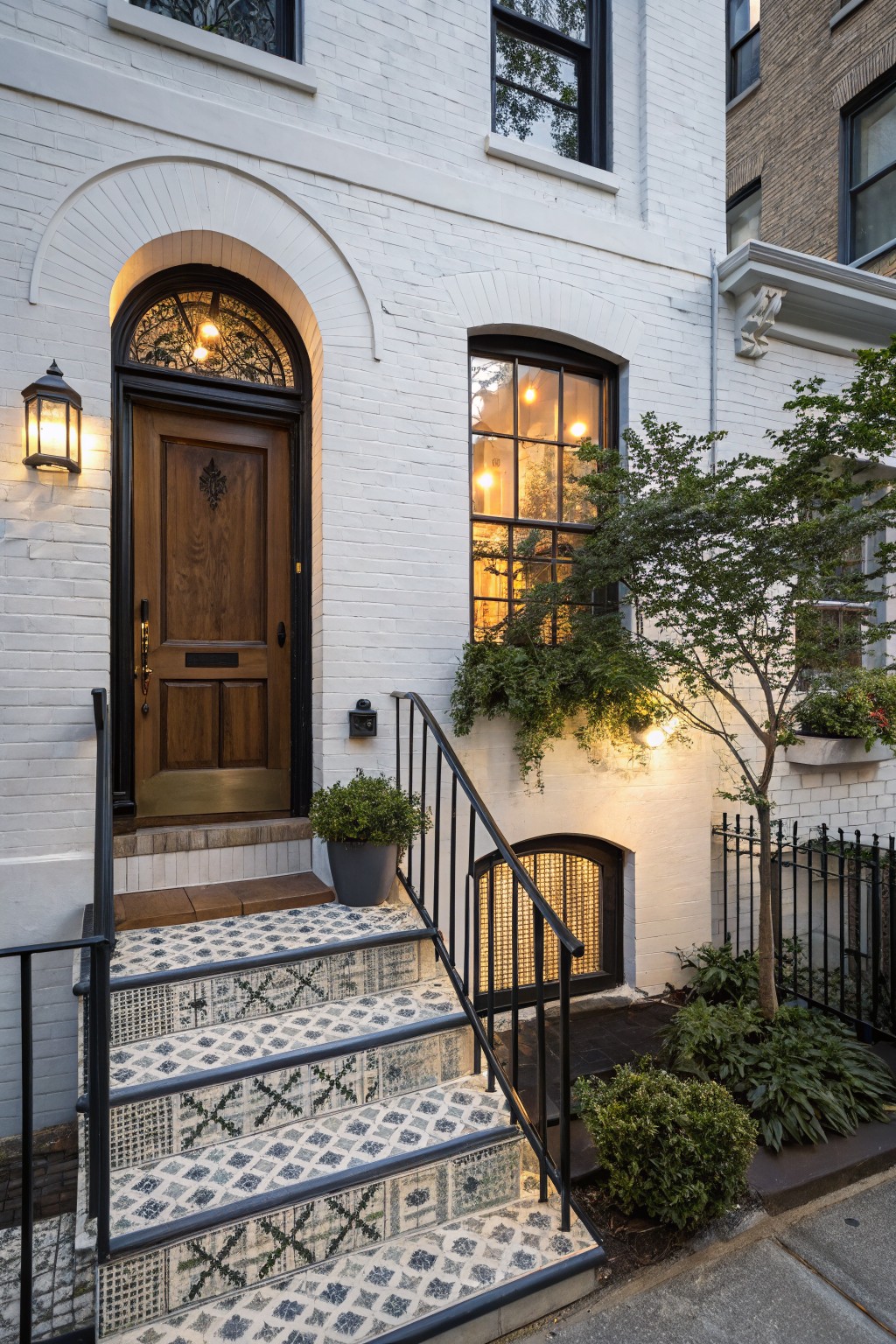 White painted brick house exterior with arched dark wooden front door, intricate blue and white patterned tile steps flanked by black metal railing, potted plants, exterior lanterns, and small trees.
