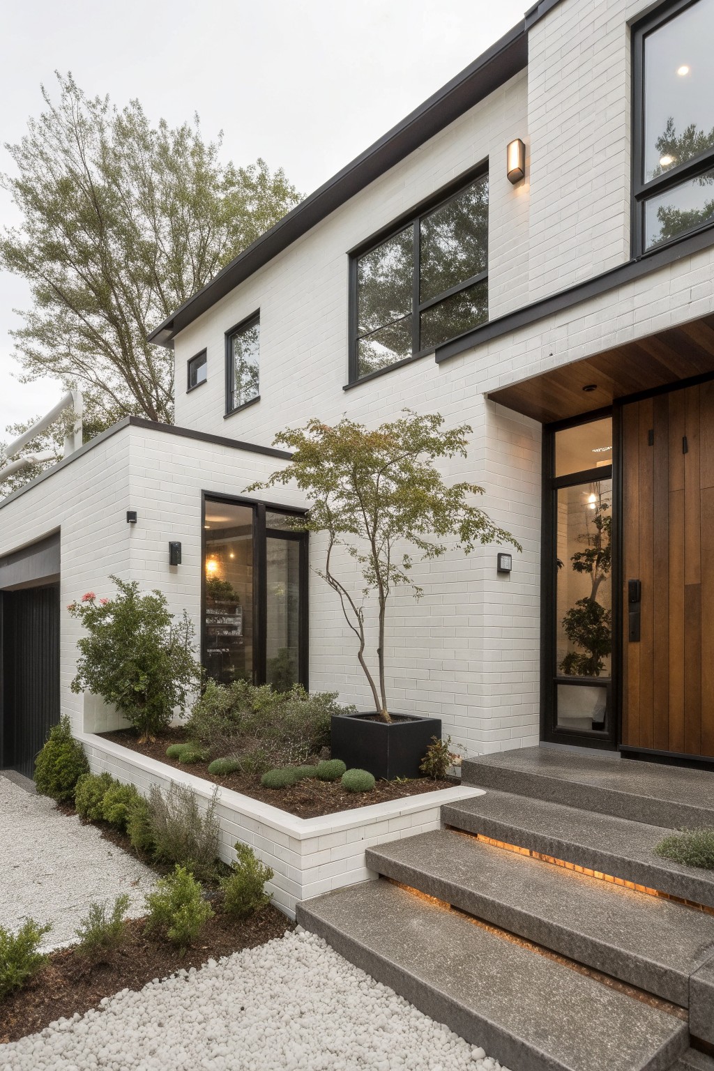 Modern white brick house exterior with black-framed windows, wooden front door, attached garage, landscaped beds with plants and trees, and concrete entry steps featuring edge lighting.