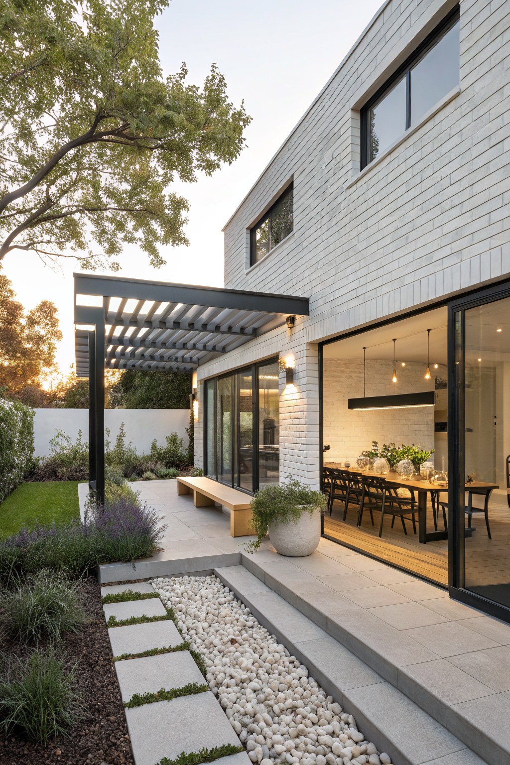 White brick house exterior with black steel pergola over concrete patio, open glass doors to indoor dining area with wooden table, stone steps leading to gravel path and garden plantings.