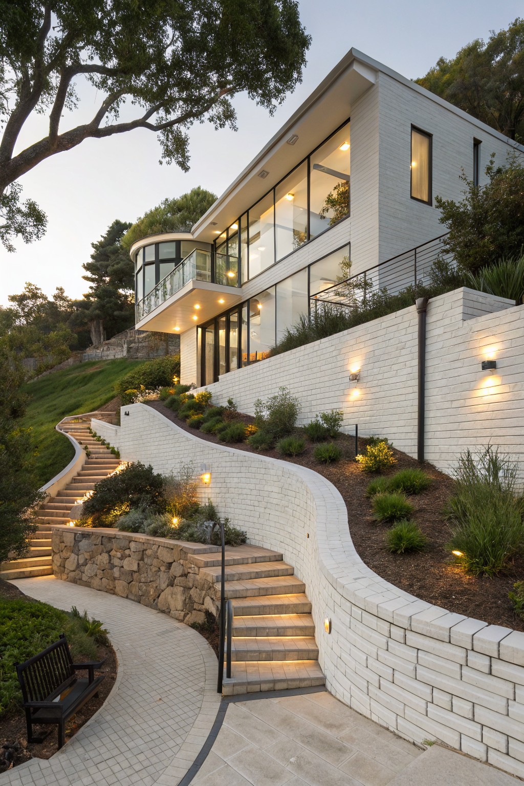 Modern white brick house on a hillside with curved matching white brick retaining walls, winding stone steps, landscaped plantings, pathway, bench, and wall lights at dusk.
