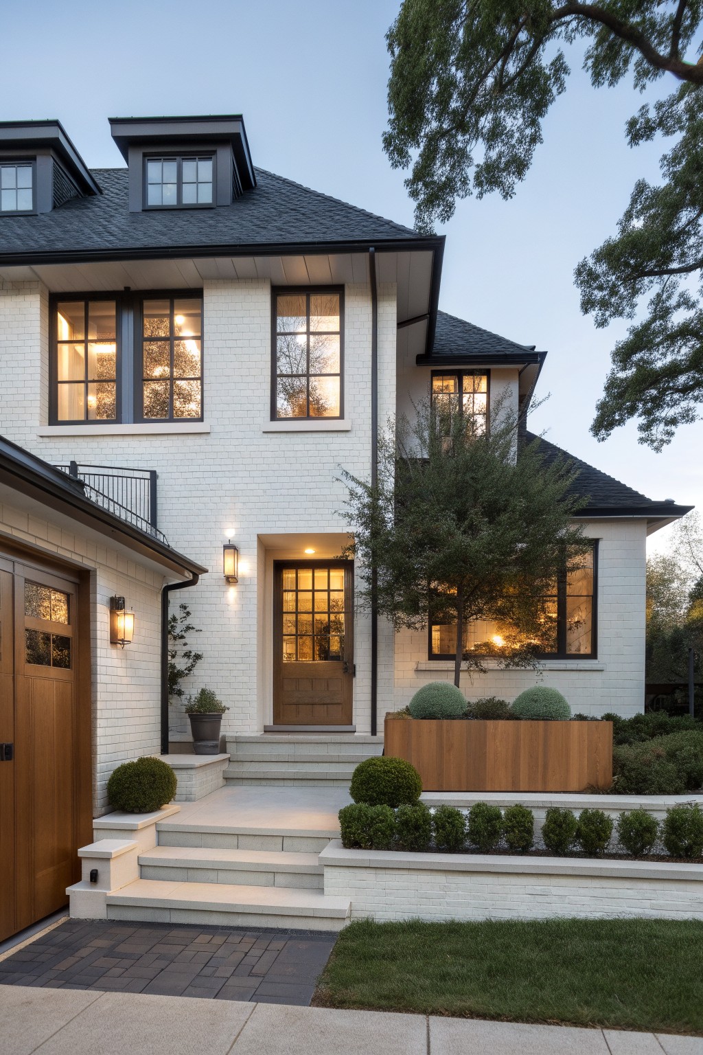 Two-story white painted brick house exterior with black roof and dormers, black-framed windows, wooden garage door, front entry steps with wood door and lantern lights, boxwood shrubs in wooden planters, and lawn at dusk.