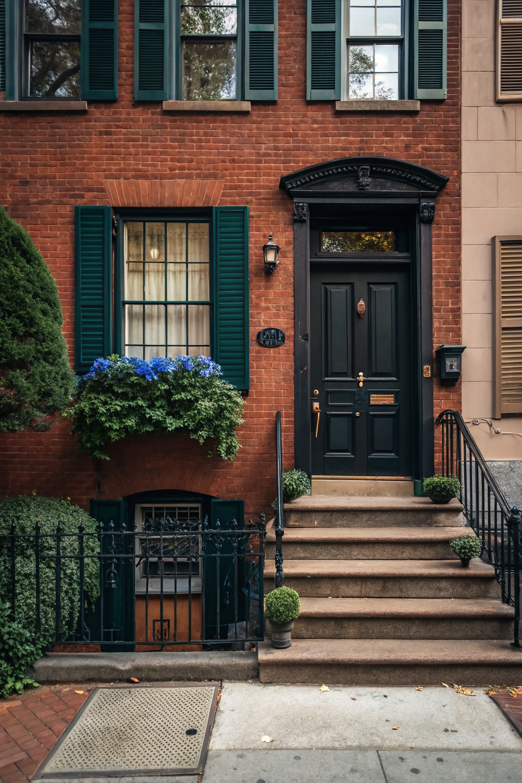 Red brick townhouse facade with hunter green shutters on windows, matte black front door with brass knocker, blue hydrangea flower box, potted plants on steps, black iron railing, and basement window grate.