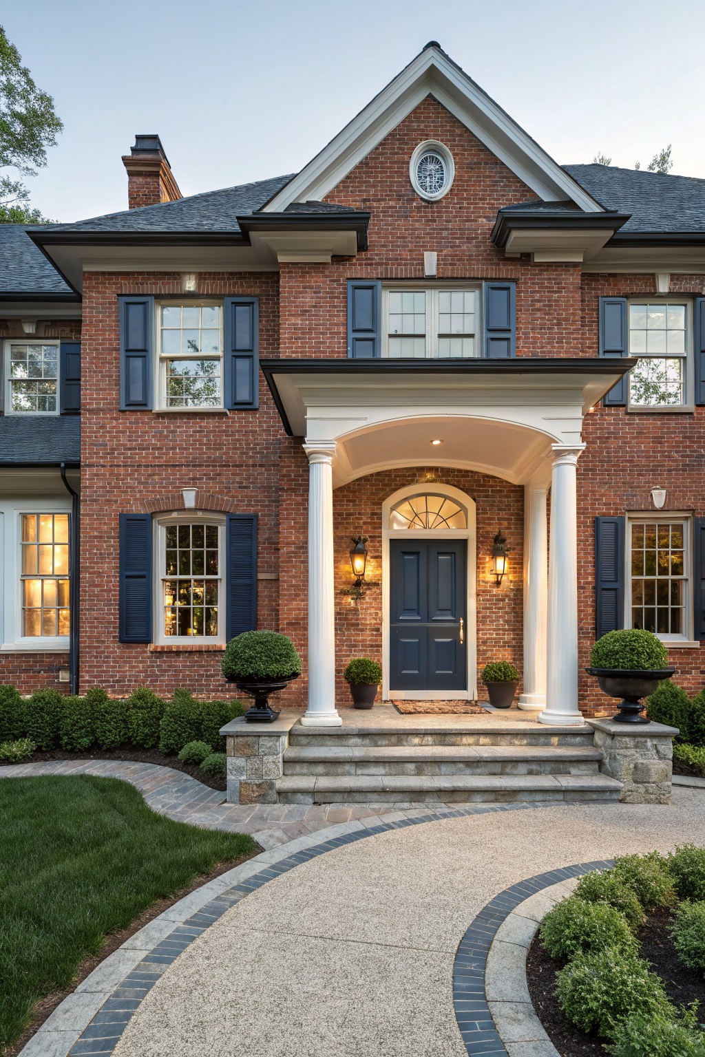 Front view of a two-story red brick house with navy blue front door and shutters, white portico and columns, lantern lights, boxwood topiaries, stone steps, and curved gravel pathway edged in brick.