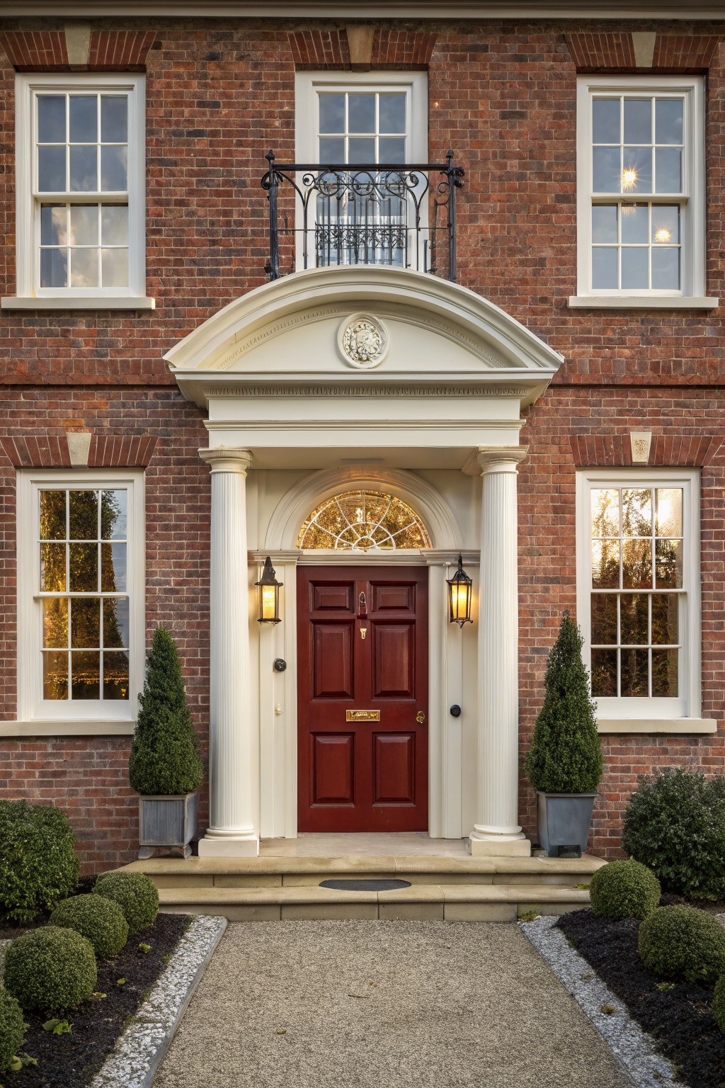 Red brick house facade with central white pedimented portico supported by columns, red front door with brass hardware, black lanterns, sash windows, boxwood topiaries in pots, and gravel path edged with plantings.