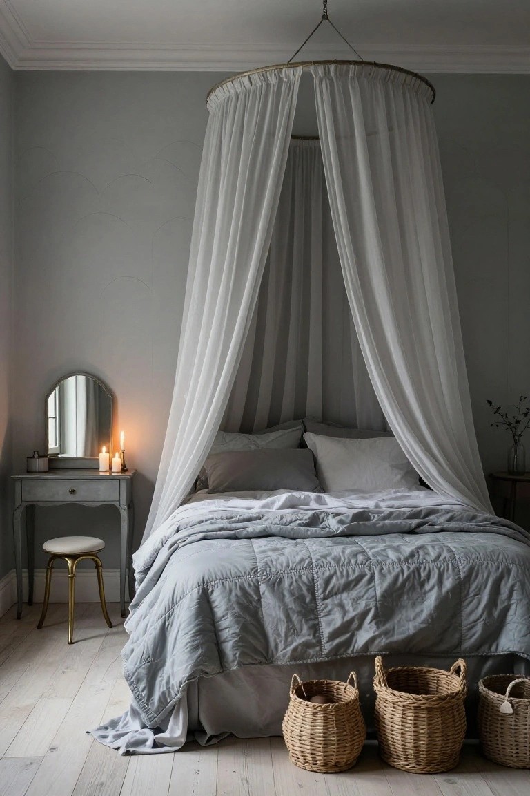 A light grey bedroom with a bed under white sheer drapes hanging from a gold hoop ceiling fixture, grey linens, a painted wood vanity table with candles and mirror, a stool, and three wicker baskets on the light wood floor.
