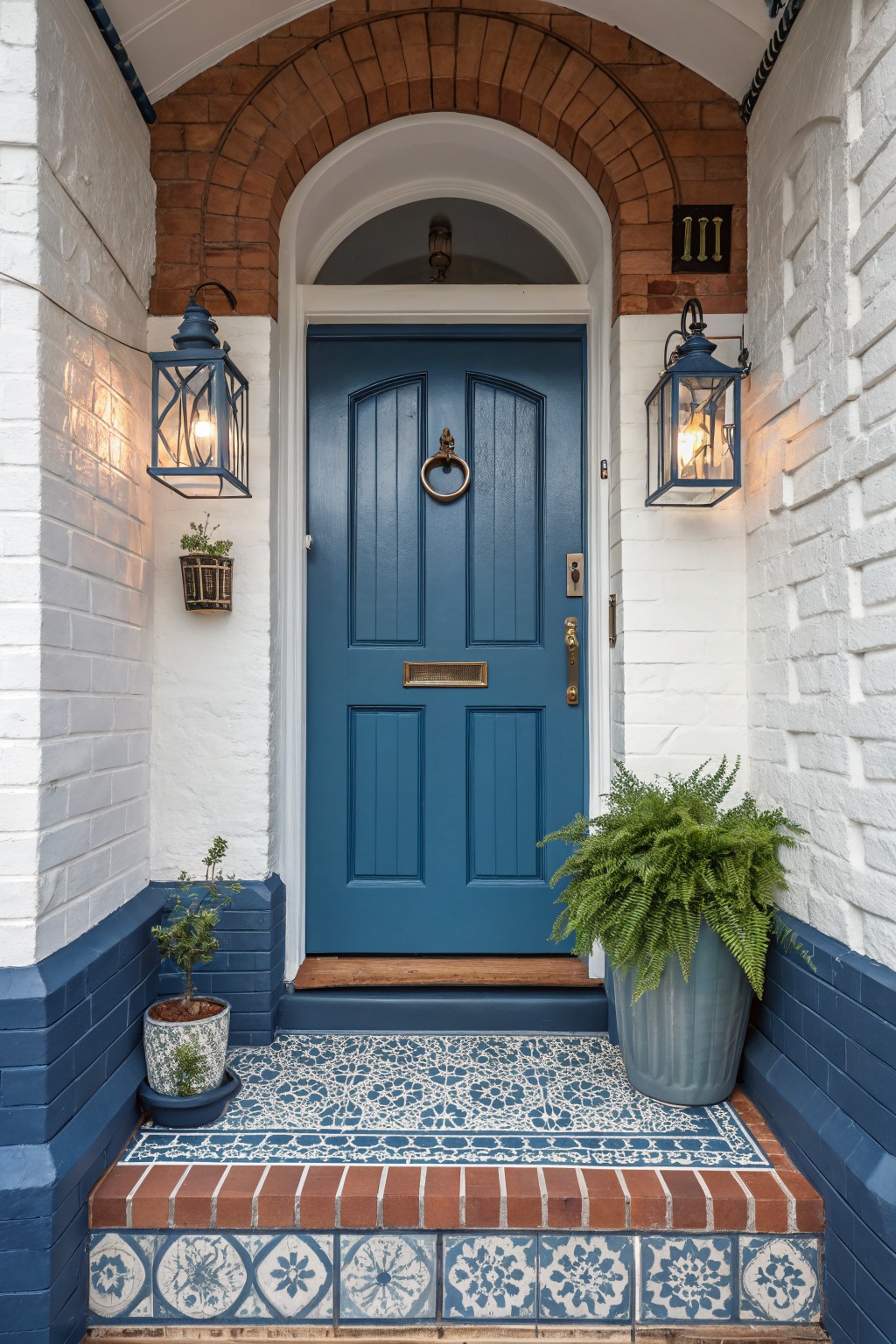 Navy blue front door under a red brick archway on white brick house exterior with blue painted base, wall lanterns, potted plants, and blue-white patterned tile doorstep.