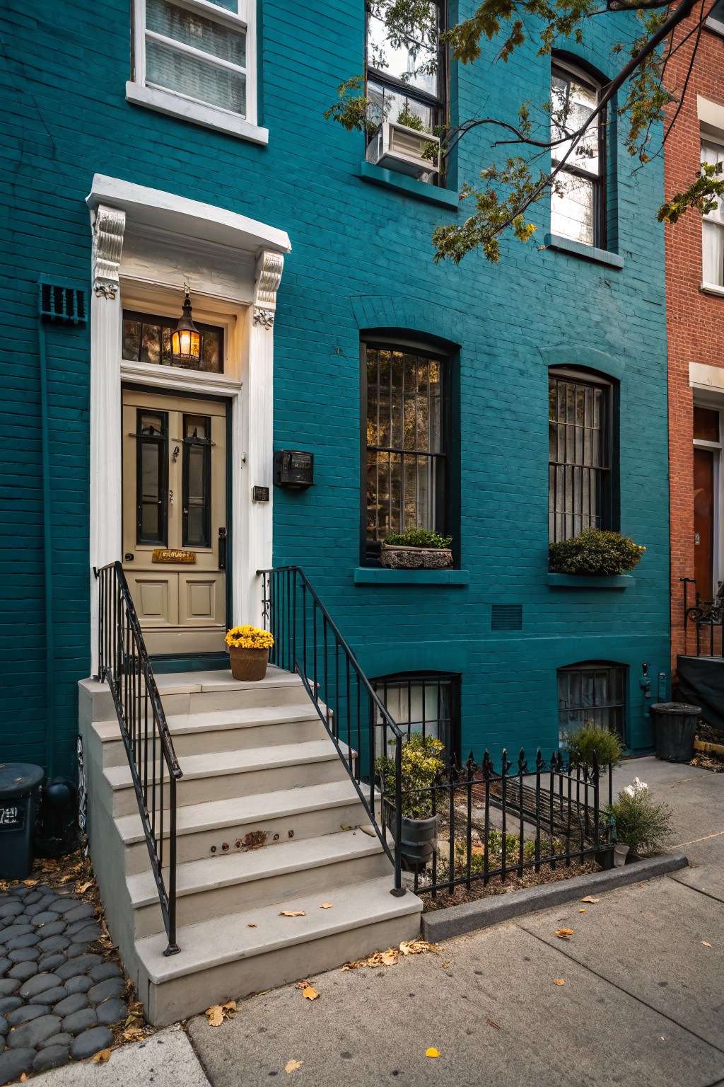 Teal painted brick rowhouse exterior with white-trimmed double doors, yellow mum flowers on front steps, black iron railing, window boxes with plants, and adjacent red brick building on a cobblestone street.