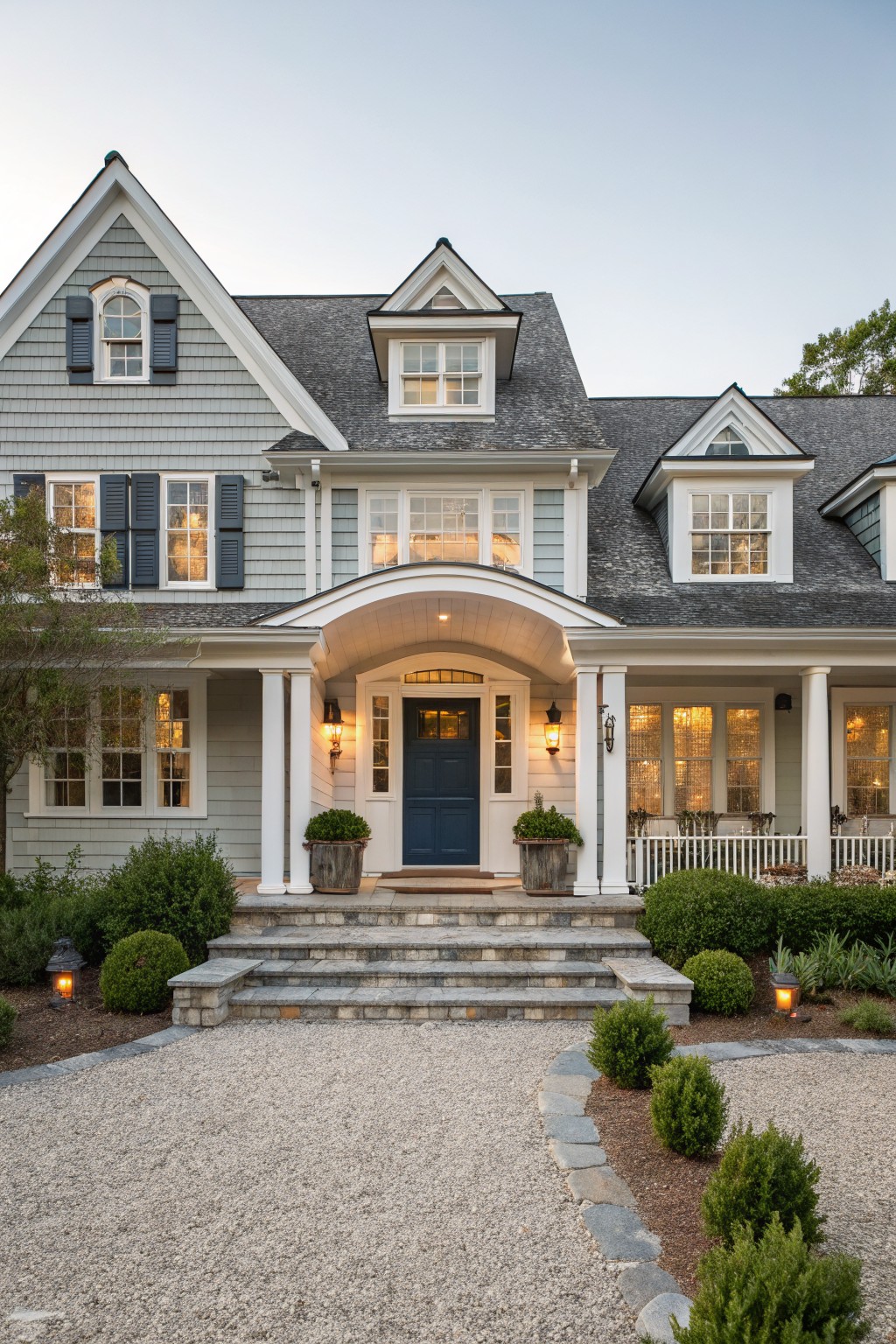 Gray shingled two-story Cape Cod house with white trim and columns supporting an arched porch over a navy blue front door, flanked by lanterns and planters, steps leading to a gravel driveway edged with shrubs.