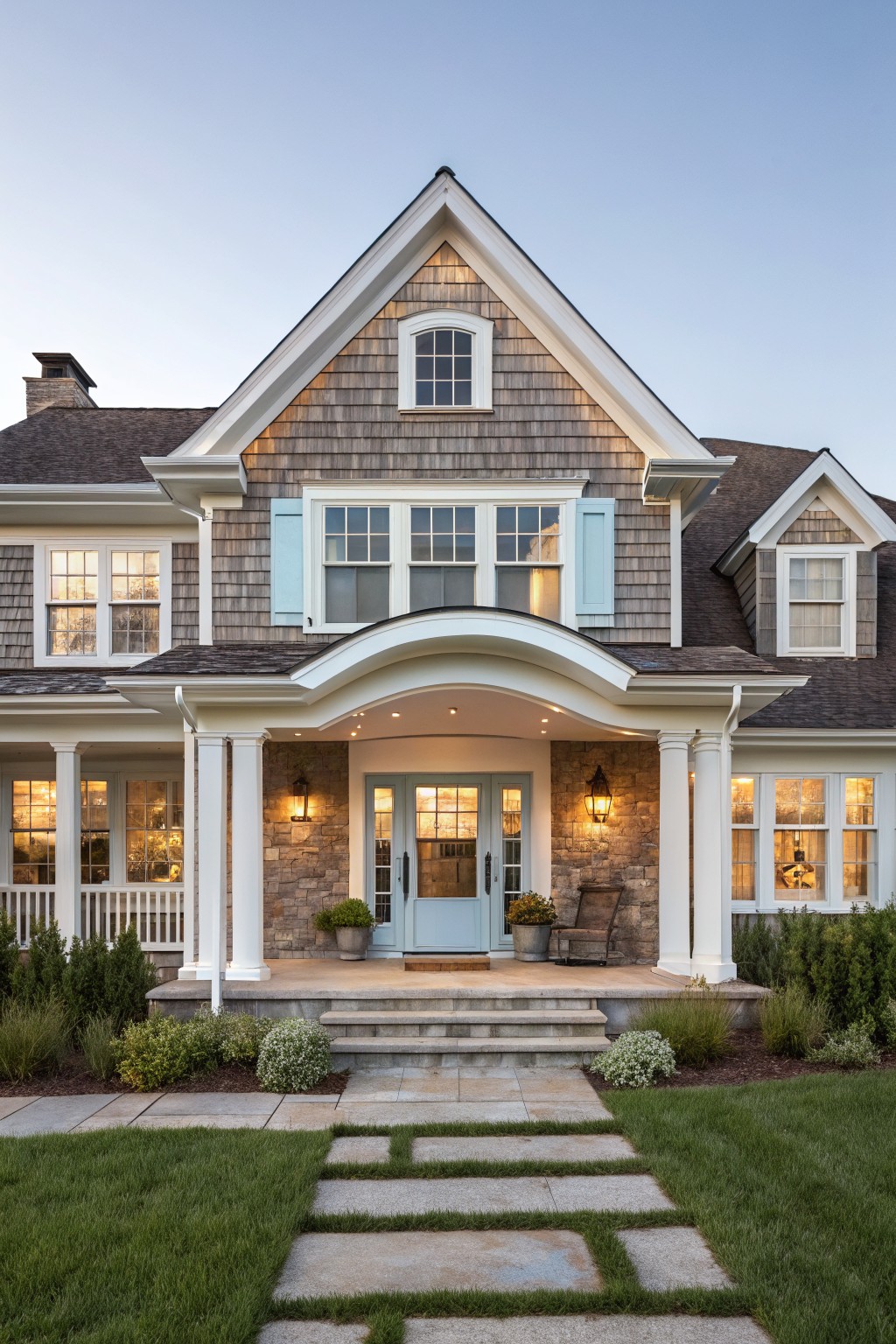Front view of a two-story shingled house with white trim, an arched portico supported by columns on stone bases, a blue front door, flanked windows, and a stone path through grass leading to steps.