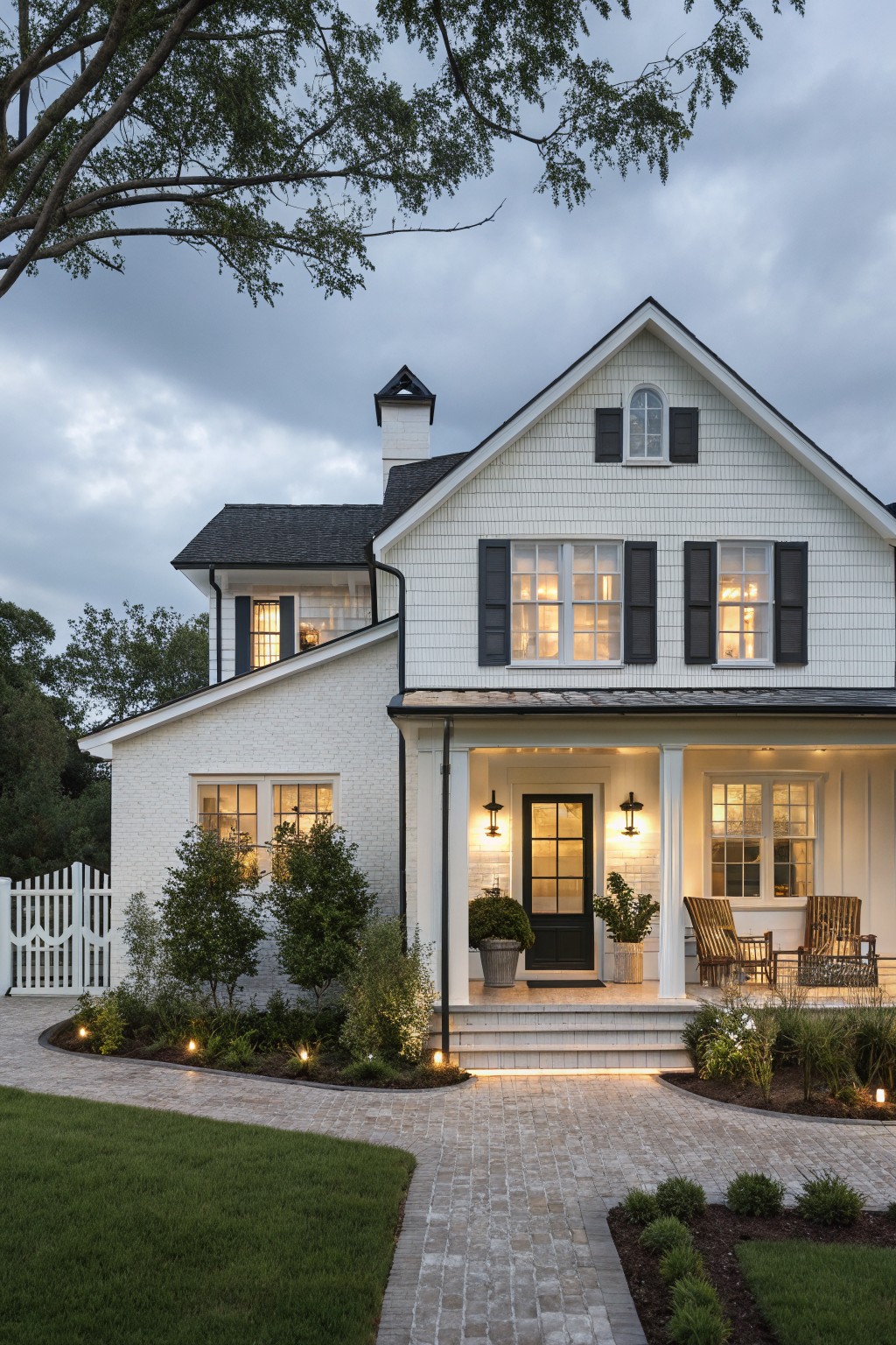 Two-story white clapboard house with black shutters, window frames, and front door under a covered porch with wicker chairs and potted plants, flanked by landscaping and a brick path leading to the entry at dusk.