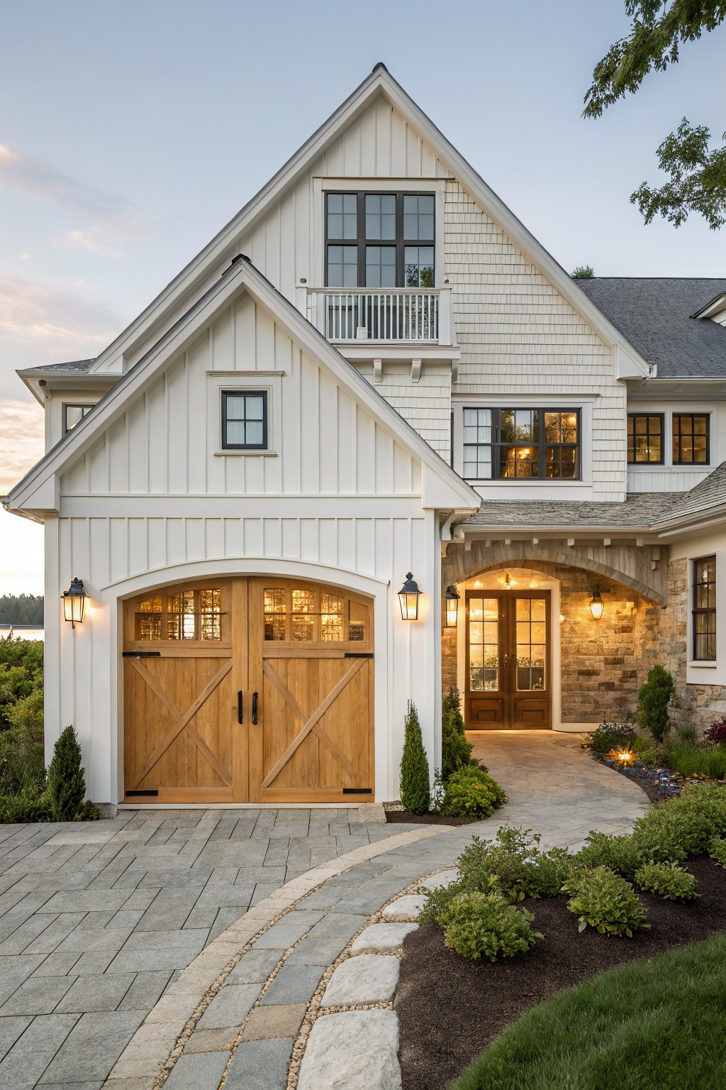 White board-and-batten sided two-car garage wing attached to a Cape Cod style house with shingle siding, wood X-braced garage doors, stone-arched entry, paver driveway curving to shrubs and grass, viewed at dusk near a lake.