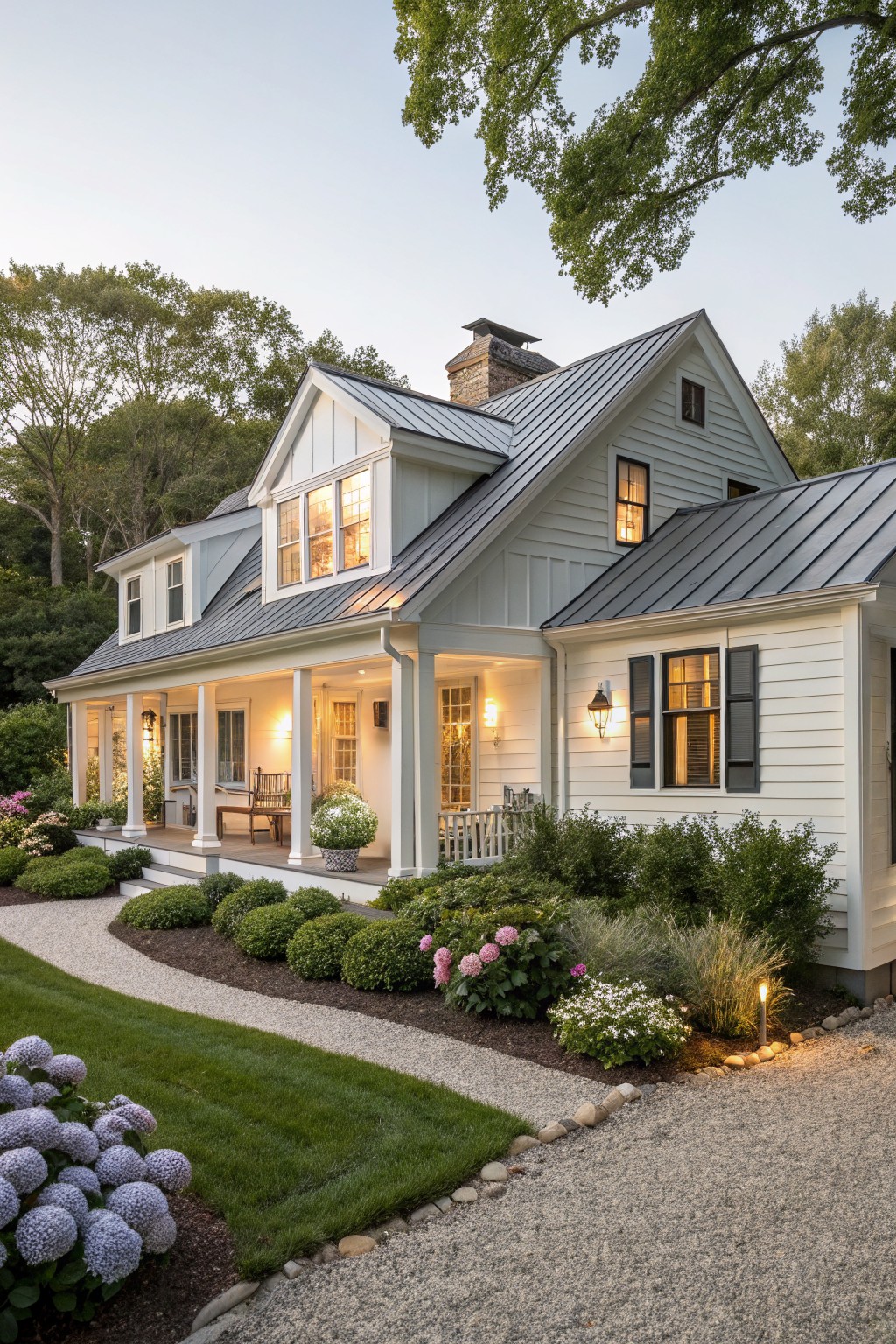 Two-story white clapboard Cape Cod-style house with dark standing-seam metal gabled roof, wrap-around porch with columns and lanterns, surrounded by hydrangeas, shrubs, gravel path, and lawn at dusk.