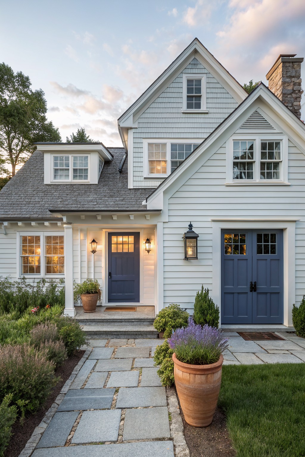 Light gray shingle-clad Cape Cod house with gabled roof, navy blue front door and double garage doors, flanked by lanterns, stone pathway, potted plants, and low shrubs.