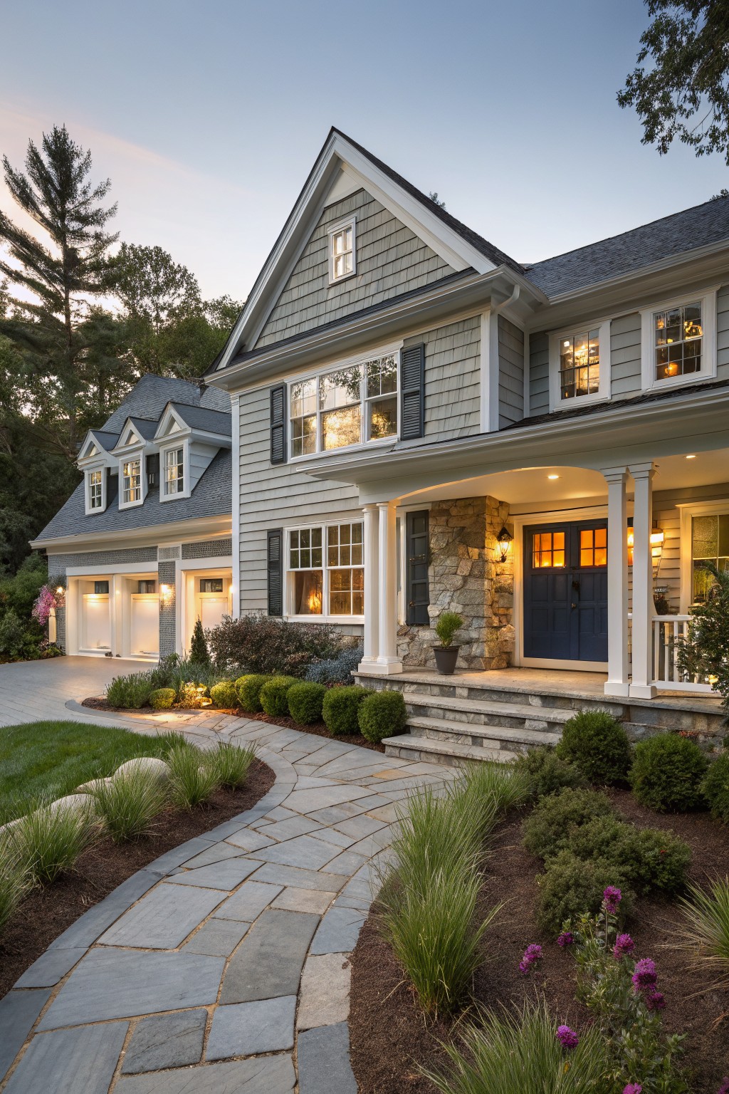 Gray shingle siding Cape Cod house exterior with attached garage, front porch supported by stone pillars and white columns, navy blue double front doors, bluestone walkway, and low plantings at dusk.