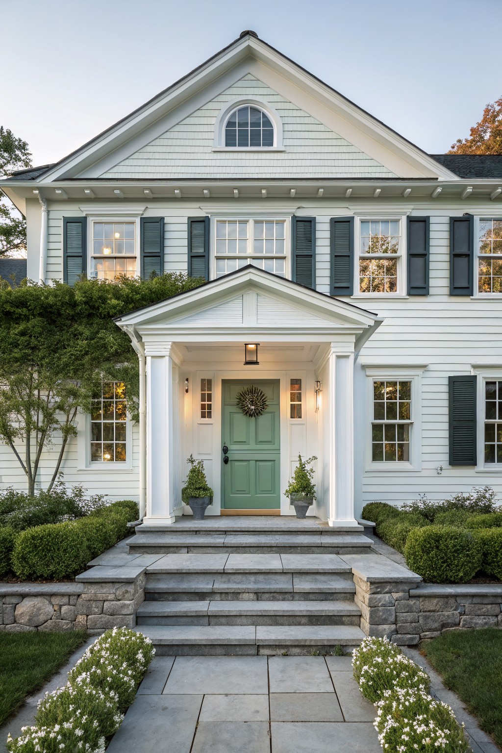 Front view of a two-story white shingle-style house with black shutters, a white pedimented portico sheltering a green double door, flanked by boxwood topiaries, stone steps, and low hedges.