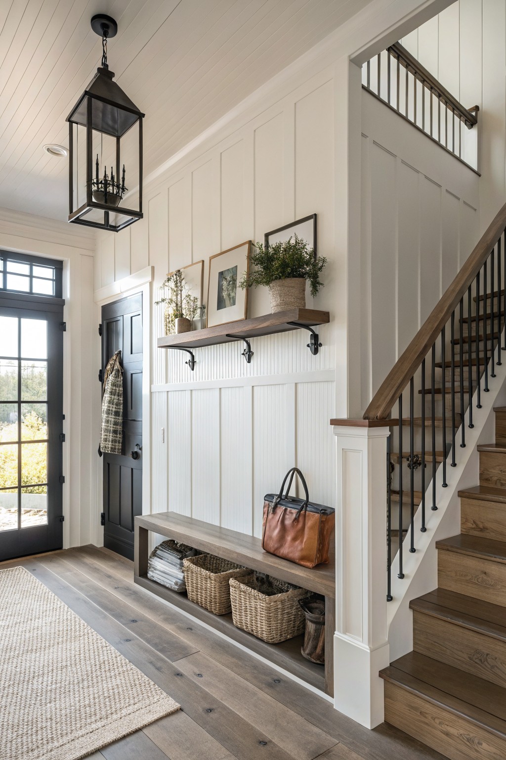 Modern farmhouse entryway interior with black front door, white shiplap walls, floating wooden shelf displaying plants and framed art, built-in wood bench with woven baskets in cubbies, and oak stairs with black railings.