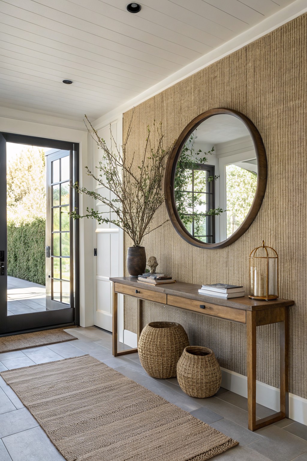Entryway with beige grasscloth wall, large round wooden mirror, wooden console table topped with vase of branches, books, and candle lantern, two woven baskets on floor, open glass door to patio, and seagrass runner on tile floor.