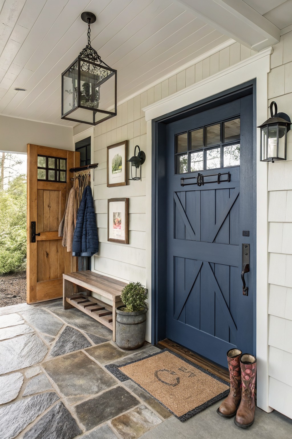 Modern farmhouse front porch entry with navy blue paneled door, open wooden storm door, wall-mounted coat hooks with hanging jackets, wooden bench with lower cubbies, potted plants in metal bucket, pair of rain boots, coir doormat, lantern light fixture, and flagstone flooring.
