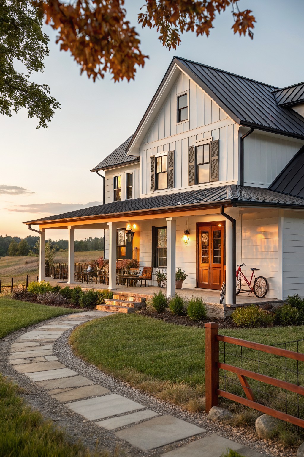 Two-story modern farmhouse with white board-and-batten siding, black standing-seam metal roof over the main house and porch, double wooden front doors, outdoor furniture on the porch, red bicycle nearby, curved stone pathway, and split-rail fence in a grassy yard with shrubs at dusk.