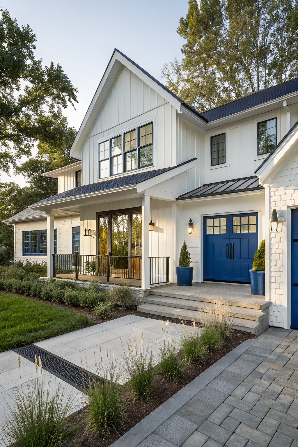 White board-and-batten modern farmhouse exterior with navy blue garage doors, black metal roof, covered porch, glass entry doors, blue window frames, steps to pathway, and surrounding landscaping with grasses and shrubs.