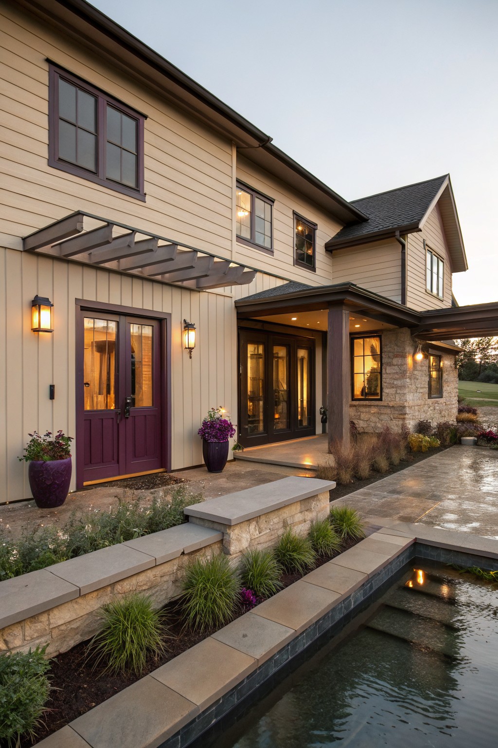 Side exterior view of a two-story modern farmhouse with beige siding, deep purple double doors flanked by lanterns and potted plants, stone accents, landscaping beds, and a nearby stone-edged reflecting pool at dusk.