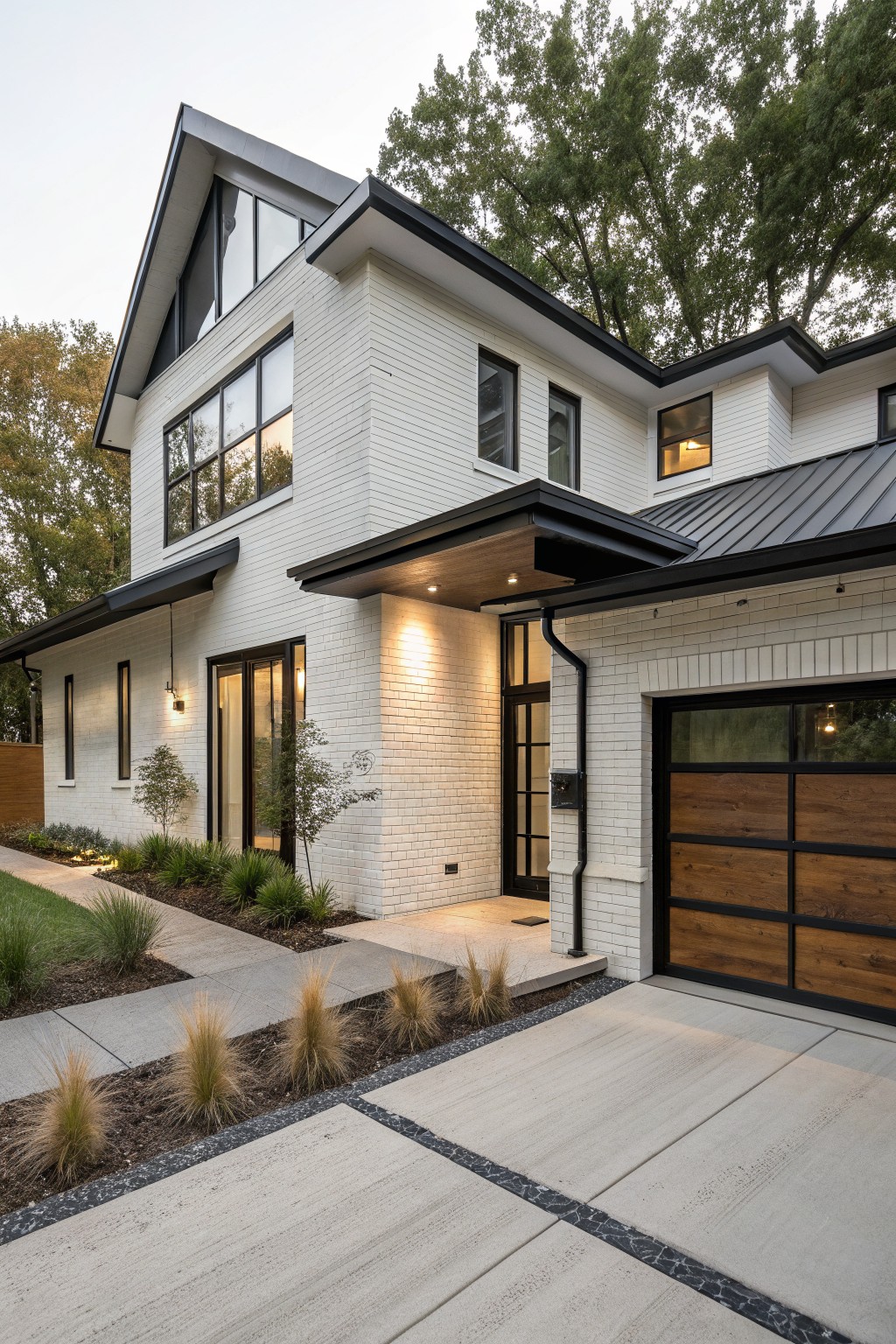 Side exterior view of a two-story white brick house with black metal roof and entry canopy, wood and glass garage door, pathway with grasses, and trees in the background.