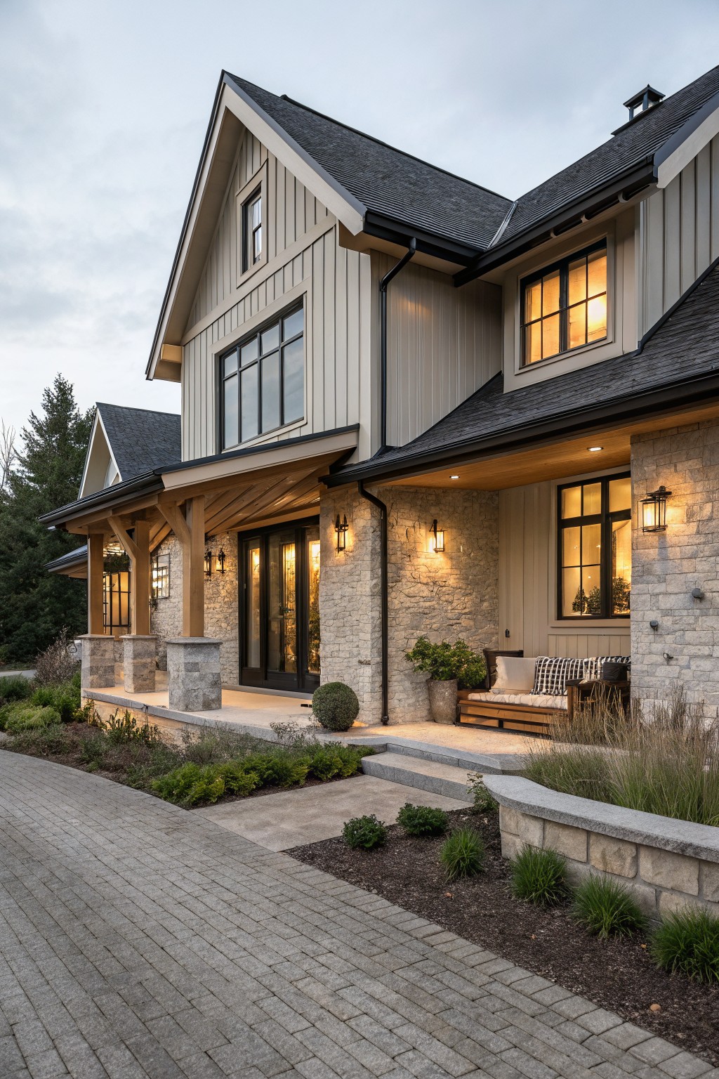 Exterior view of a two-story modern farmhouse house with light gray board-and-batten siding, stone pillar supports on a covered front porch, black-framed windows and doors, warm lantern lighting, and surrounding landscaping at dusk.