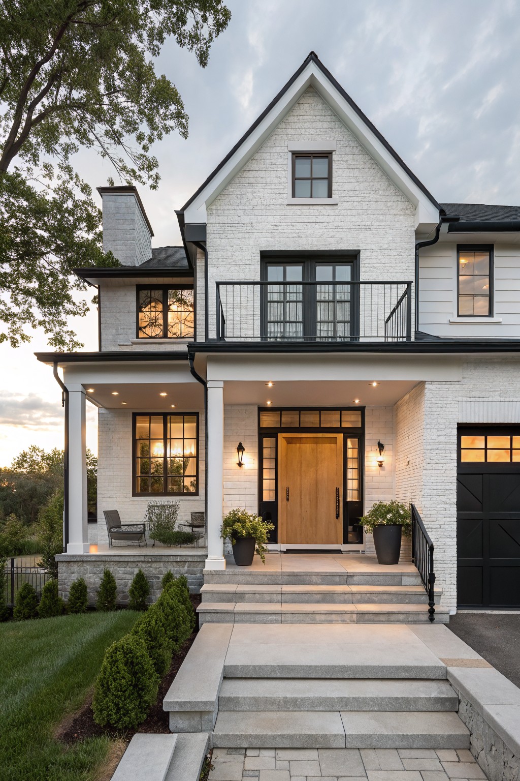 A two-story white brick house with black roof, trim, windows, garage door, covered porch with columns, wooden front door, stone steps, and surrounding landscaping and trees at dusk.