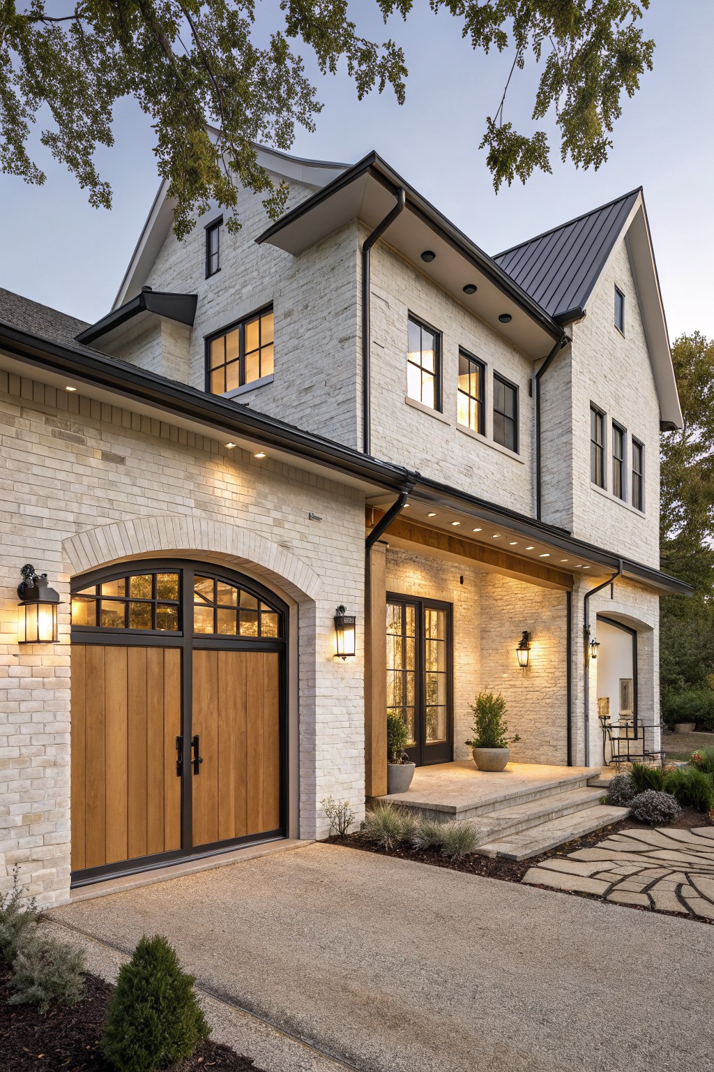 Two-story modern farmhouse exterior with white brick walls, black metal standing seam roof, large arched double wood garage doors flanked by lanterns, entry porch with glass door, concrete driveway, stone steps, low shrubs, and landscape lighting at dusk.