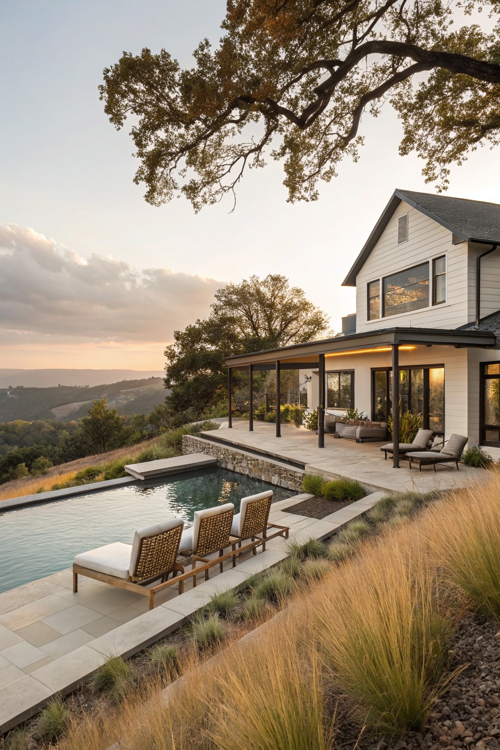 White modern farmhouse with gabled roof and large windows, black metal pergola-covered terrace with three woven lounge chairs, rectangular pool on stone deck overhanging grassy hillside toward valley landscape at sunset, oak trees in foreground.