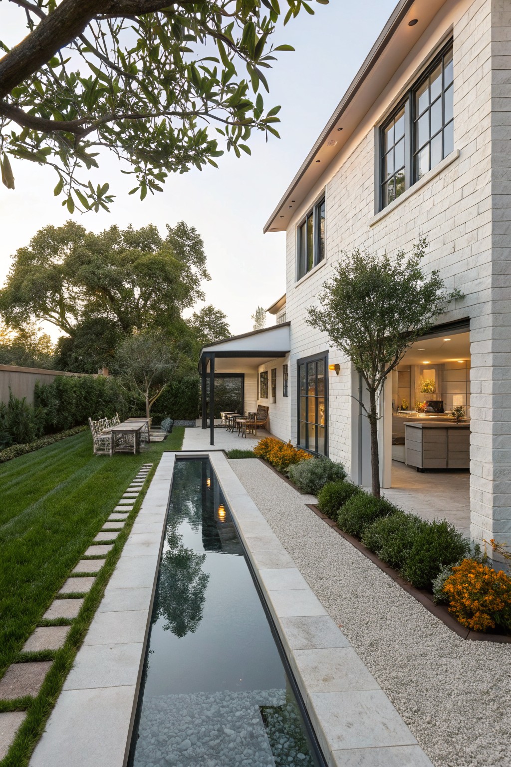 Side view of a white brick modern house with a long narrow reflecting pool pathway edged in gravel and stepping stones, leading from a grassy lawn past trees and plants to an open outdoor kitchen and dining area under a pergola.