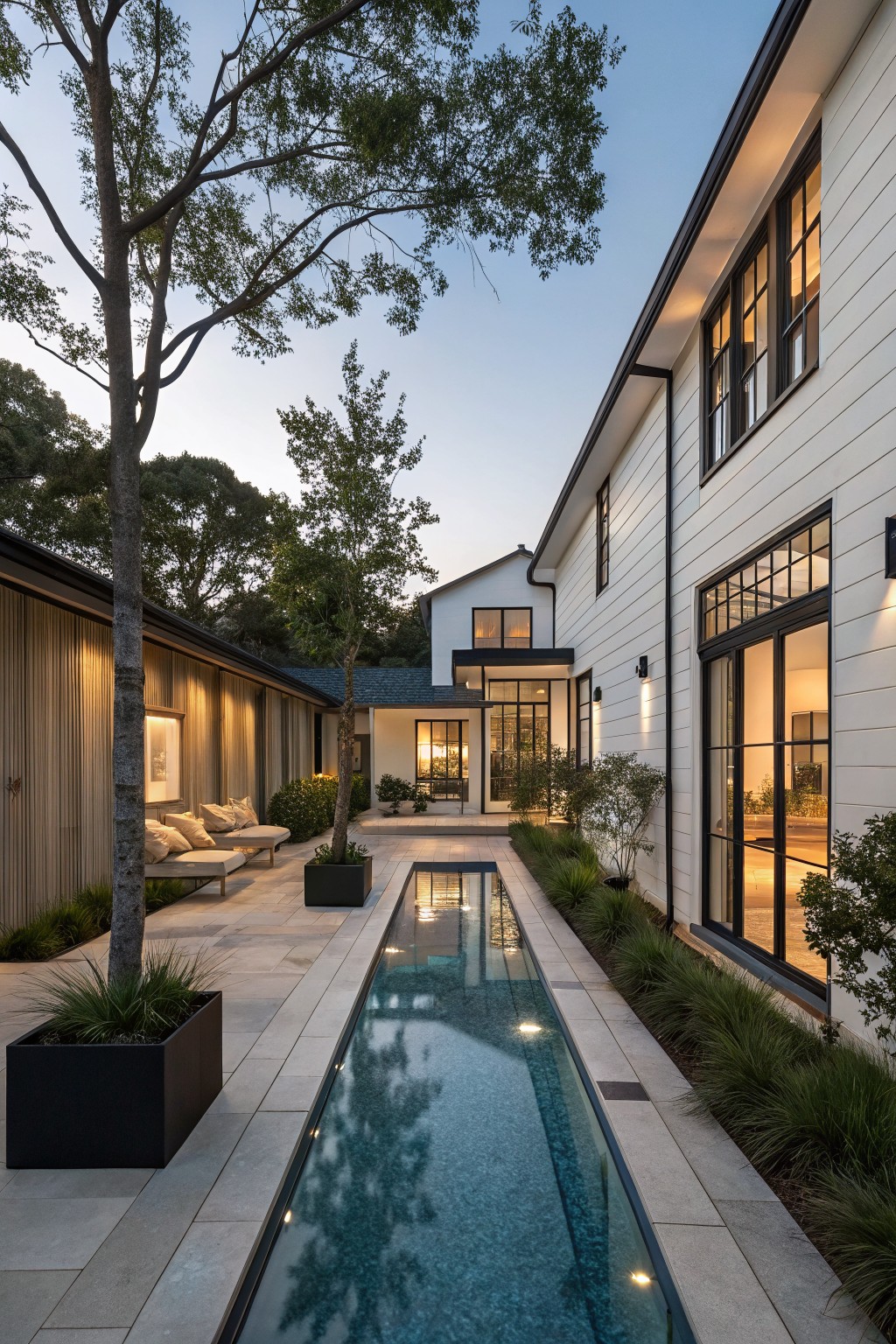 White modern farmhouse with black-trimmed windows and a long narrow lit pool running parallel to the side wall, surrounded by trees, plants in black planters, and beige outdoor loungers.