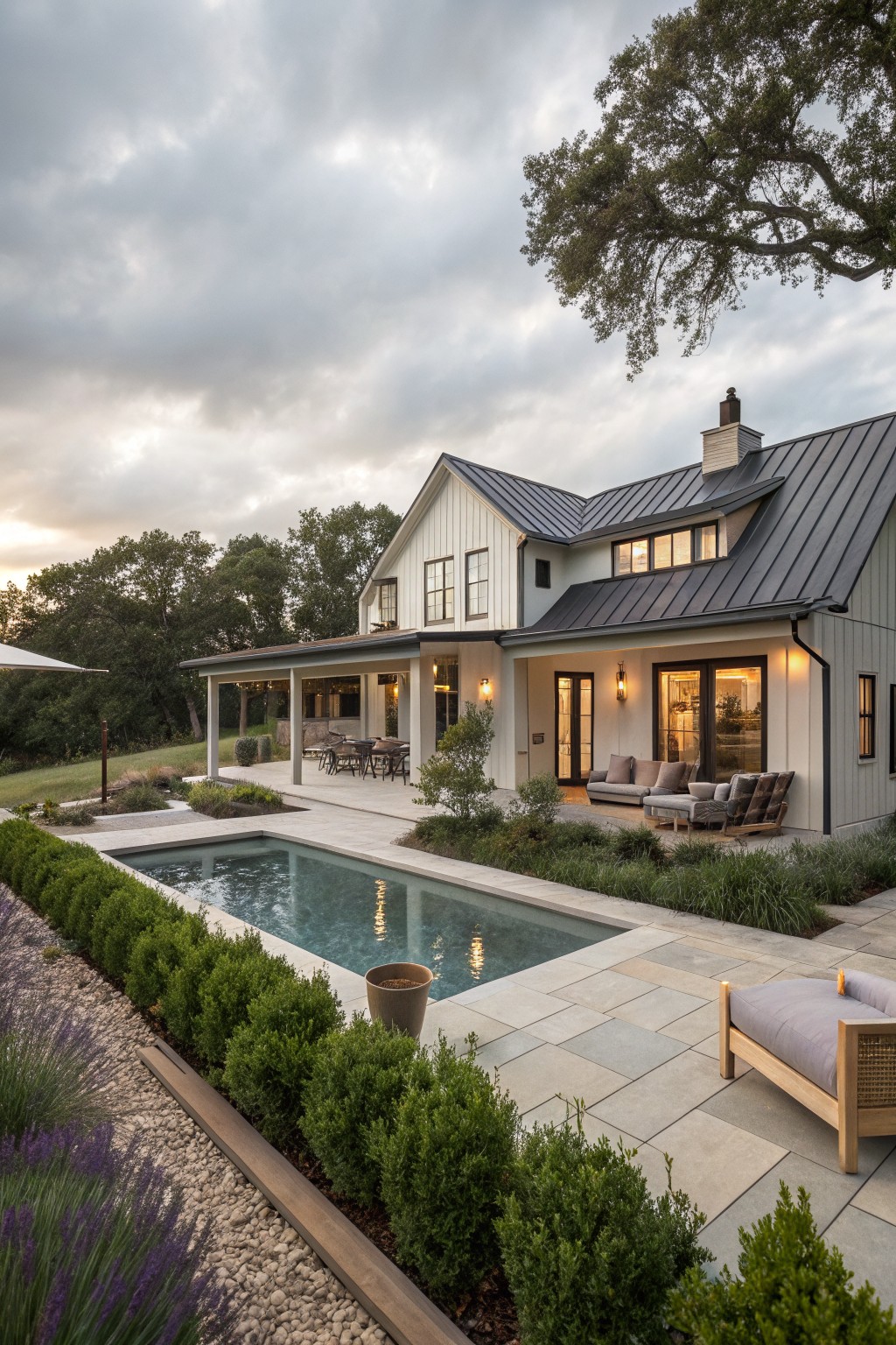 Back exterior of a modern farmhouse with white board-and-batten siding, dark metal gable roof, covered porch with lounge seating, rectangular pool, boxwood hedges, lavender plants, and gravel edging on a patio.