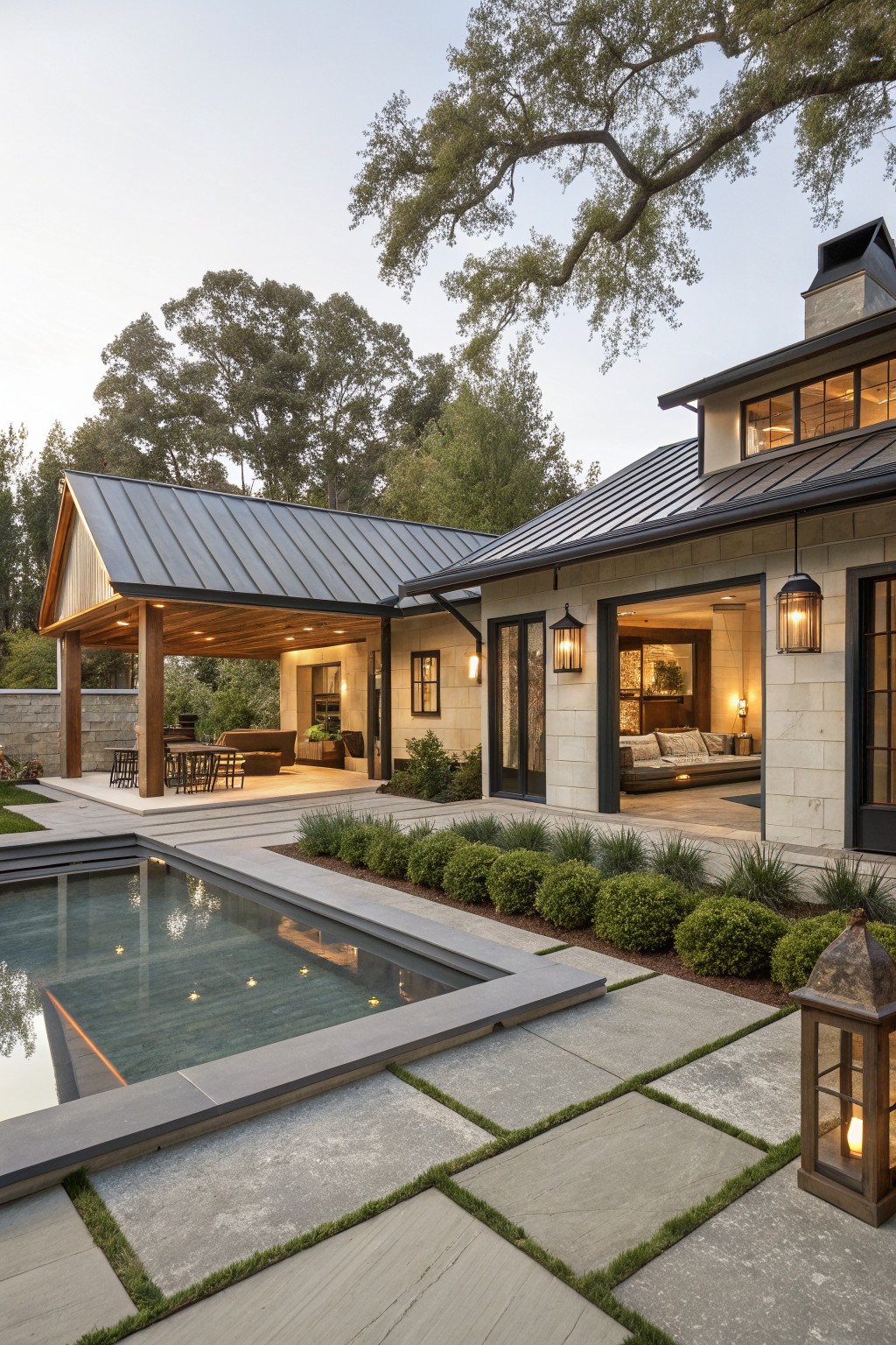 Rear view of a modern stone house with a covered porch featuring wood beams and metal roof, large open garage doors showing interior seating, adjacent rectangular pool with dark water, paver patio, boxwood shrubs, and lanterns.