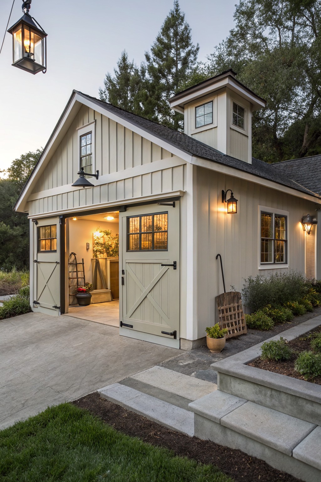 Detached two-car garage with light beige board-and-batten siding, open cross-braced doors revealing lit interior with plants and tools, small cupola on roof with window, wall-mounted lanterns lit, driveway apron, steps, and landscaped beds with trees at dusk.