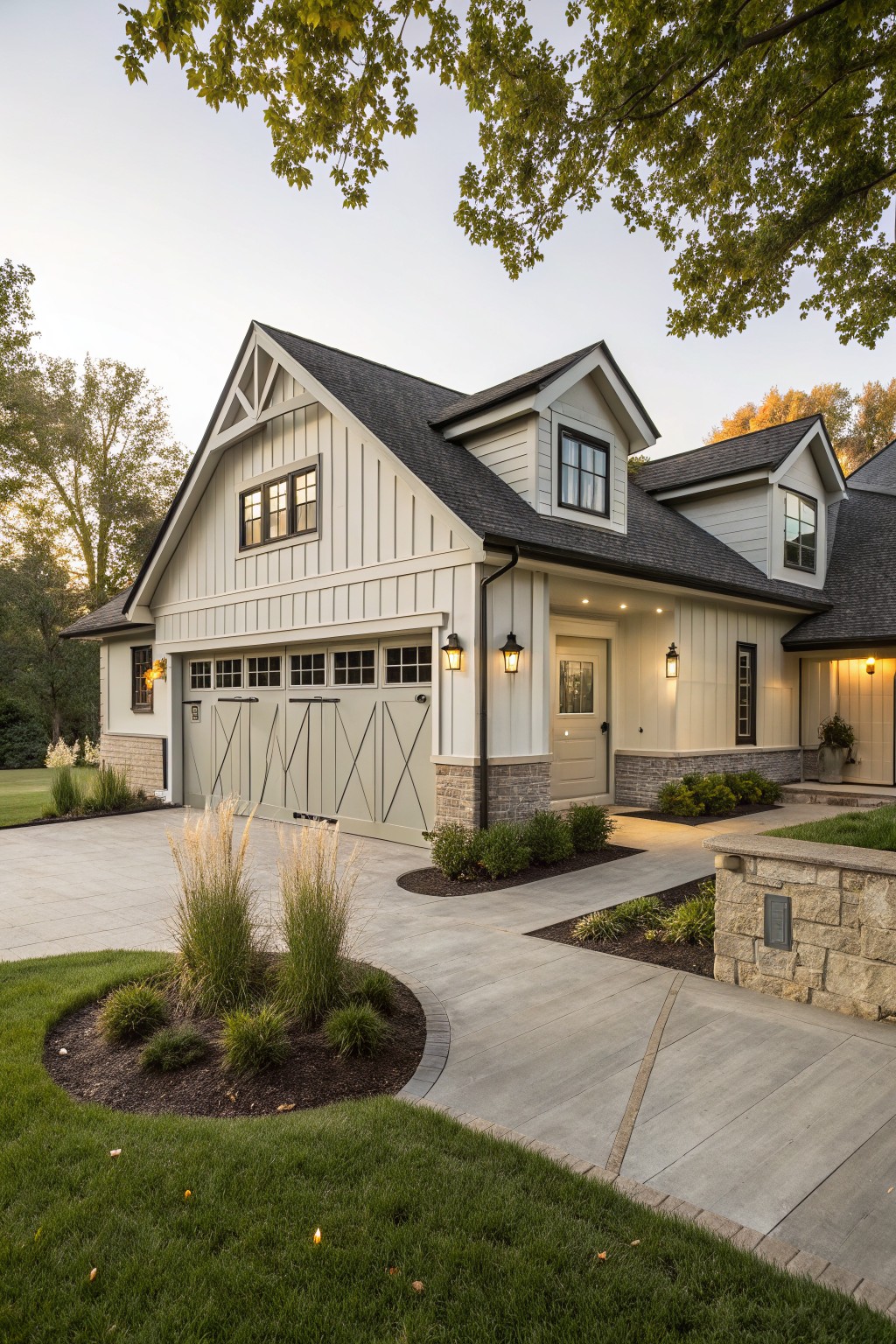 Exterior view of a modern farmhouse house with white board-and-batten siding, attached two-car garage featuring gray X-braced doors, curved concrete driveway with central landscaping island of grasses and shrubs, walkway to side entry door, and lantern lights.