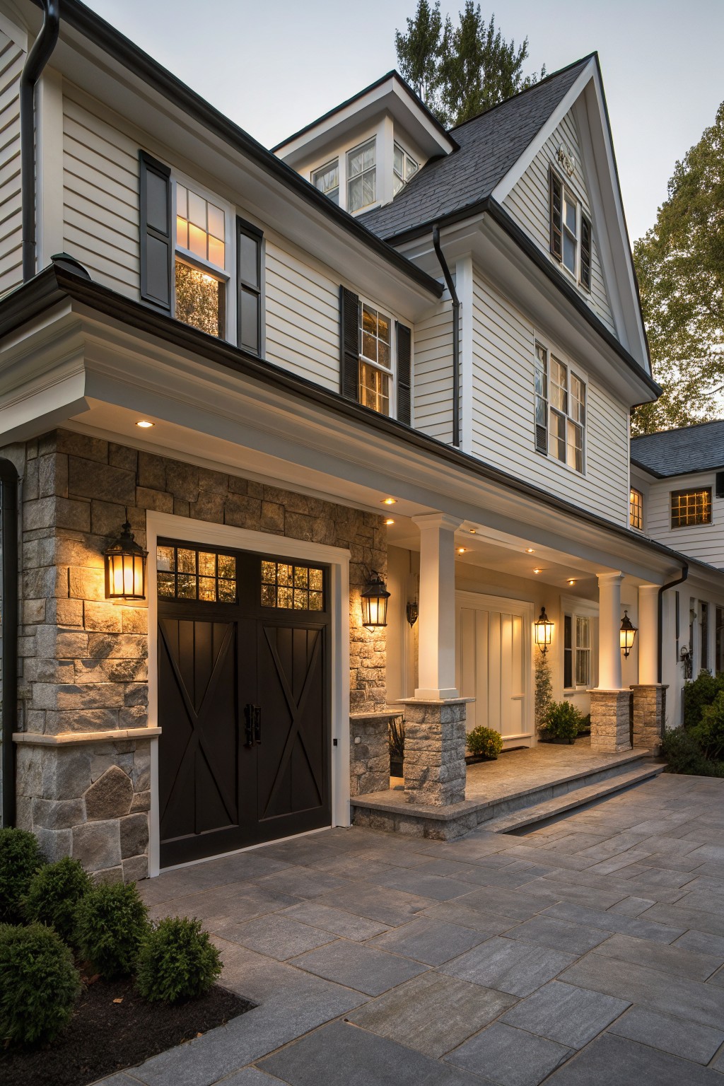 Exterior of a two-story modern farmhouse home with white clapboard siding, dark double garage doors surrounded by stone walls and pillars, lanterns on either side, covered porch with columns, and stone paver driveway.