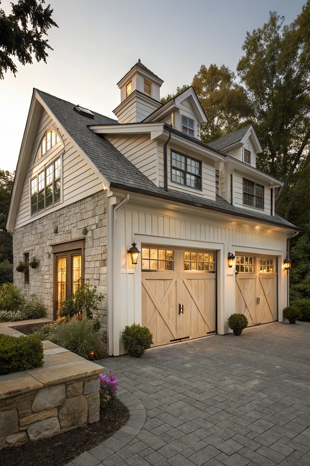 Two-car garage with light wood plank doors, stone foundation, white board-and-batten siding, large upper-level windows, and a cupola on a shingled roof, next to a paver driveway and landscaping.