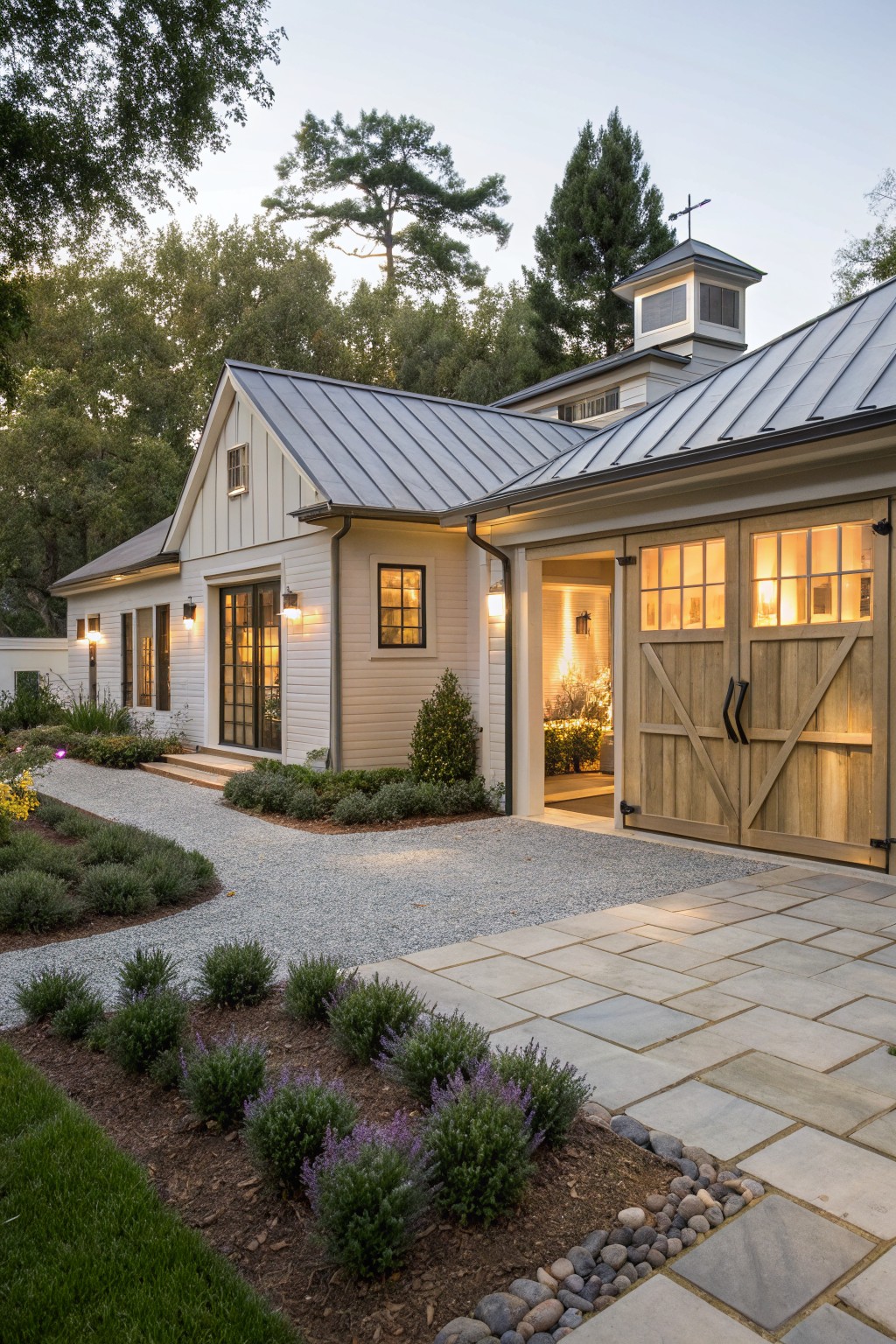 Side view of a modern farmhouse house with attached open garage featuring double wooden doors, metal roof extending over a breezeway area, gravel path, stone pavers, and surrounding shrubs and trees at dusk.