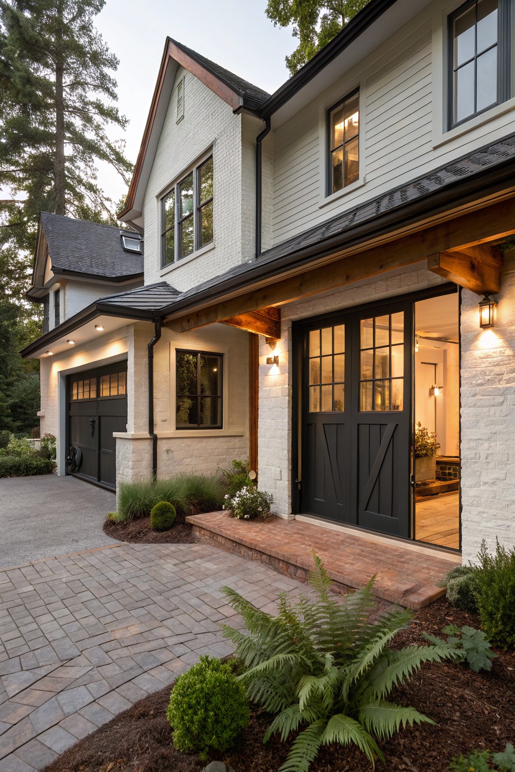 White modern farmhouse house exterior with side-facing black garage doors, open glass entry doors under wooden porch beams, brick paver driveway curving to the garage, stone accents, and low plants along the front at dusk.
