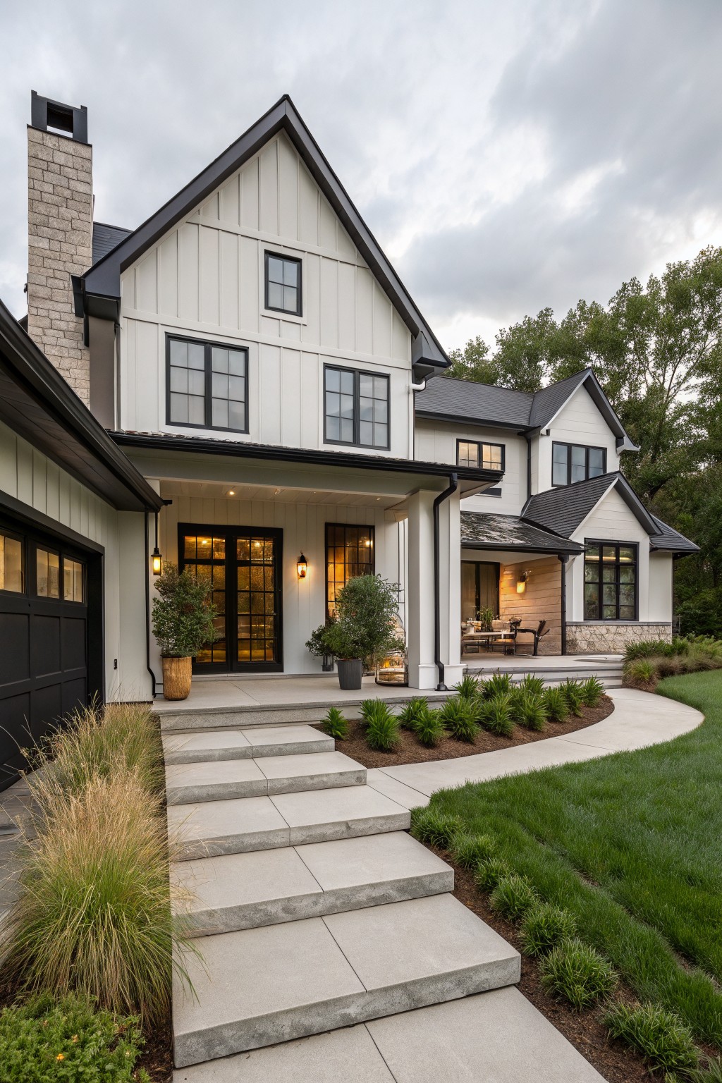 Two-story modern farmhouse house exterior with white board-and-batten siding, black window frames and trim, stone chimney, attached black garage door, covered front porch with glass doors, concrete steps, and low landscaping along a curved path.