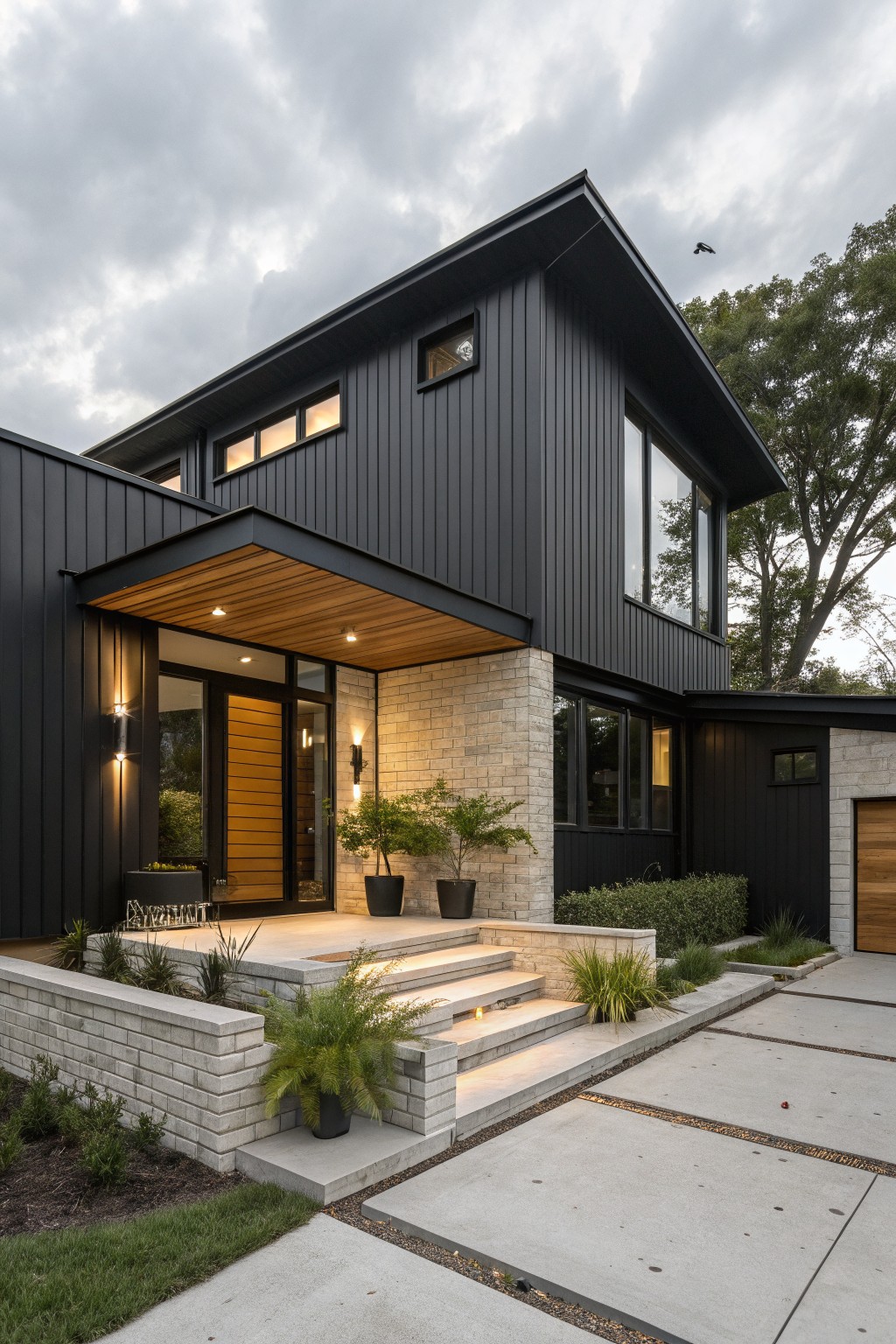 Modern two-story house exterior featuring black vertical siding on upper levels, light brick base, wooden canopy over the entry door, steps with lighting, potted plants, and surrounding low landscaping on a concrete pathway.