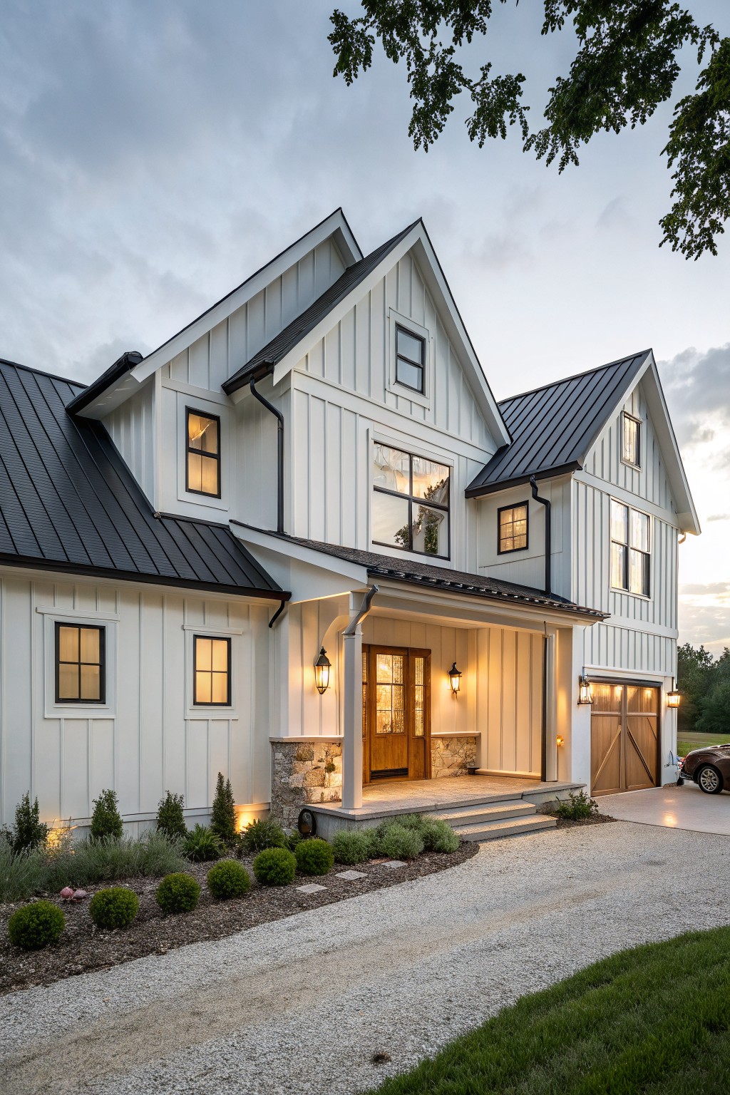 Two-story modern farmhouse house with white vertical board-and-batten siding, black metal roof, covered front porch, wooden garage doors, stone accents, gravel driveway, and low shrubs.