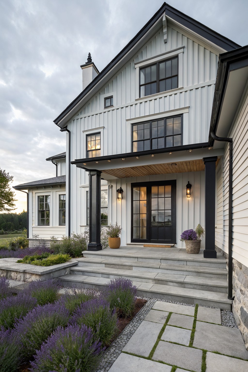A two-story modern farmhouse exterior with light vertical board-and-batten siding, black-trimmed windows and gabled roof, covered entry porch with wood ceiling and blue door, concrete steps, and lavender plants in the front yard.