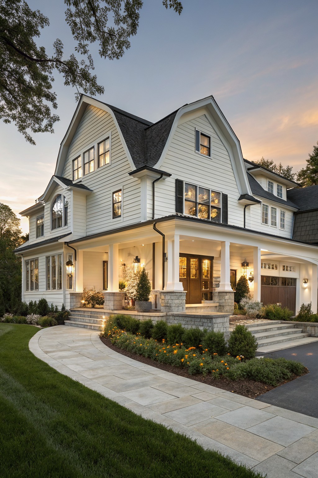 Three-quarter view of a two-story modern farmhouse with white clapboard siding, dark gray gabled roof, wraparound front porch, attached garage, stone entry steps, curved paver path, and landscaped borders at twilight.
