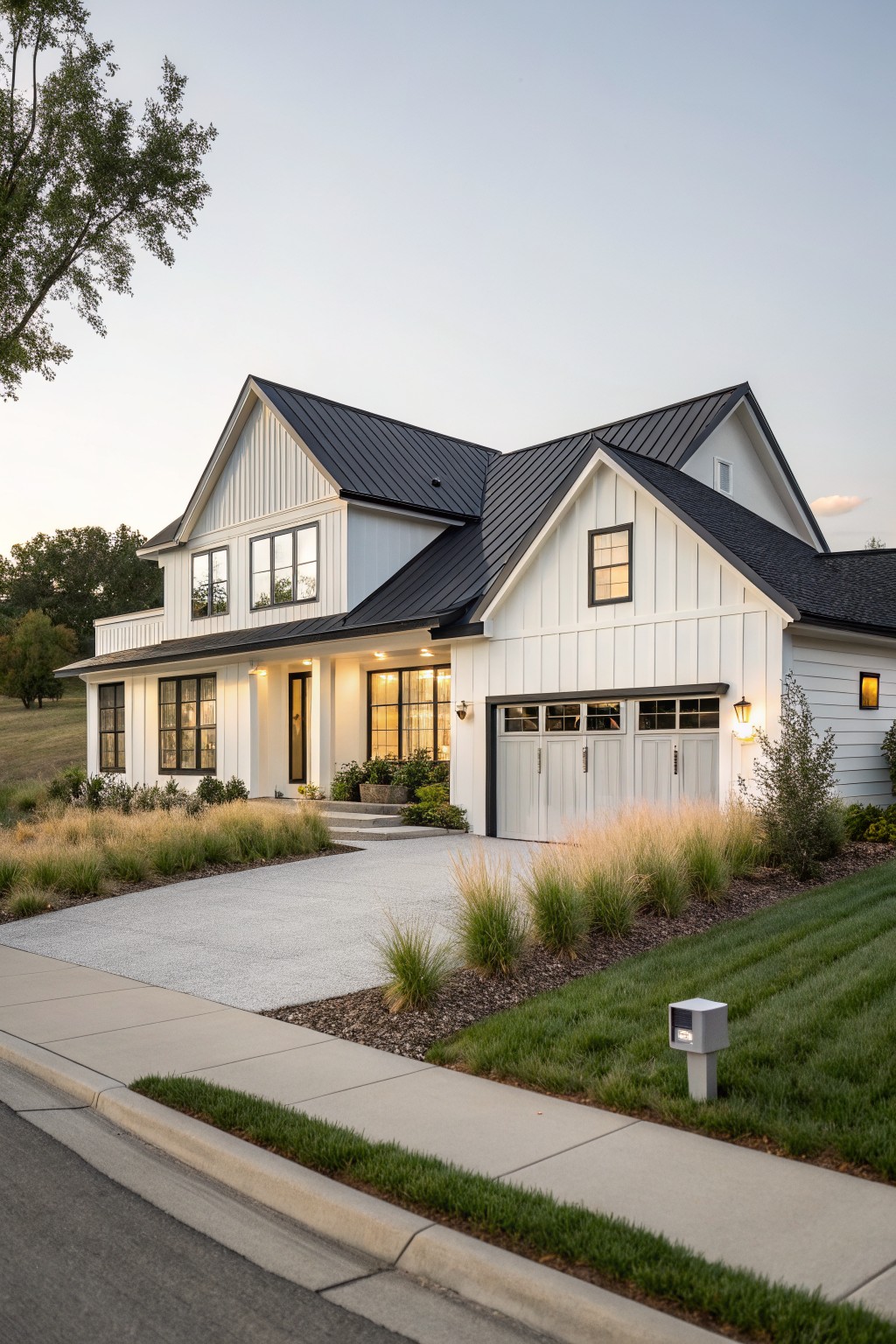 White board-and-batten sided modern farmhouse house with black metal gable roof, attached two-car garage with gray doors, concrete driveway, ornamental grasses, lawn, and street curb at dusk.