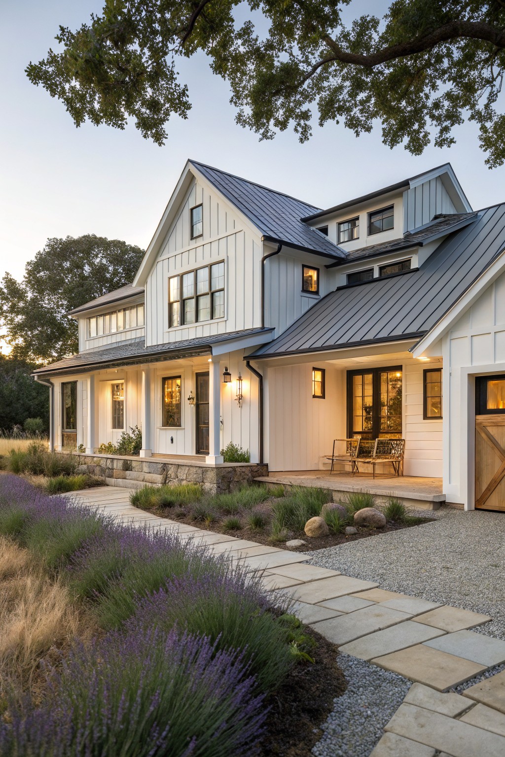 Exterior of a single-story modern farmhouse with white board-and-batten siding, dark standing-seam metal roof, covered porch with lanterns, barn-style garage door, and gravel pathway bordered by lavender plants and grasses.